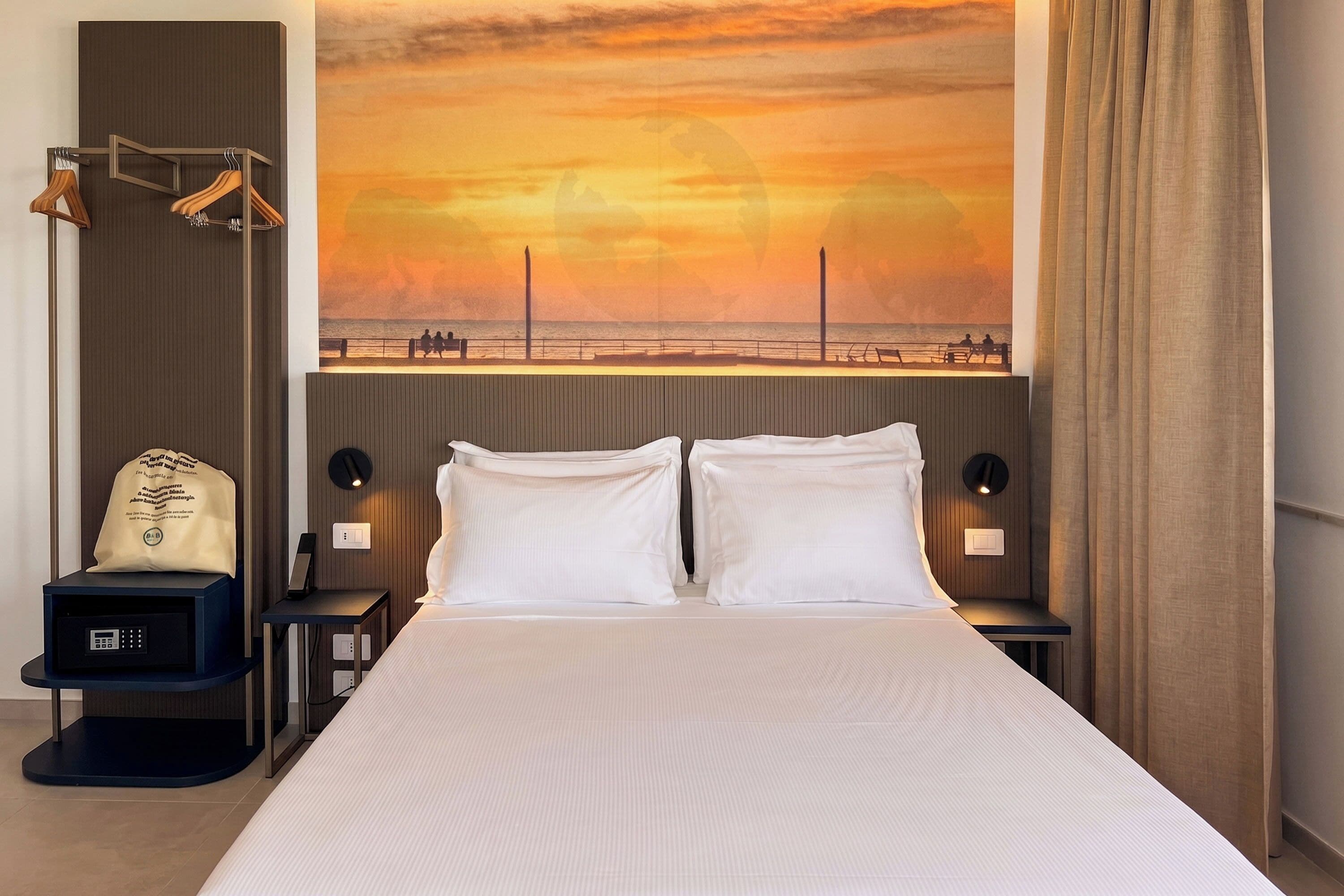 Une chambre d’hôtel avec un grand lit, deux tables de chevet, un porte-manteau fixé au mur, une horloge numérique et un grand tableau représentant un coucher de soleil.