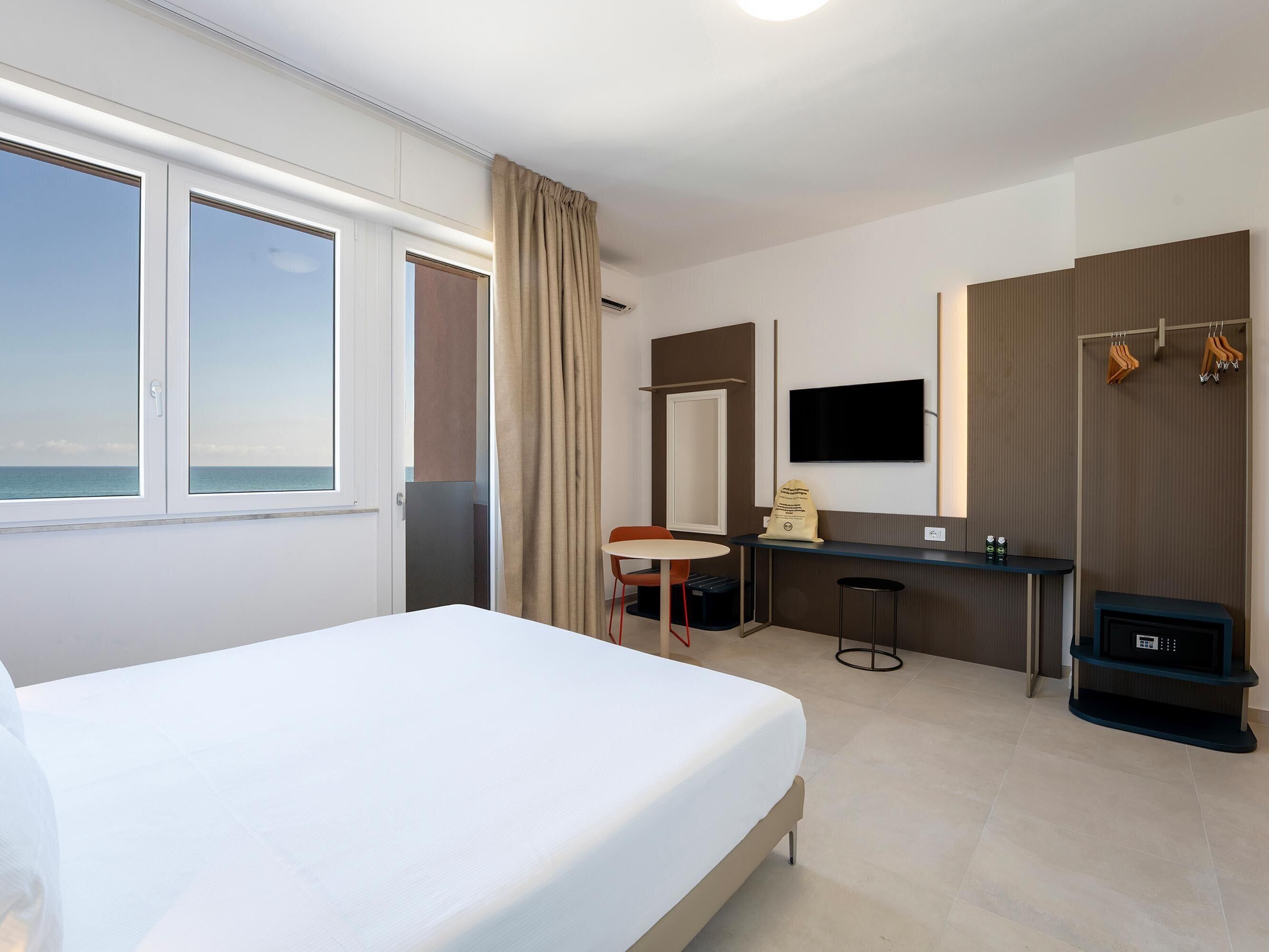 A hotel room with a bed, a desk, a chair, a TV, and a view of the sea.