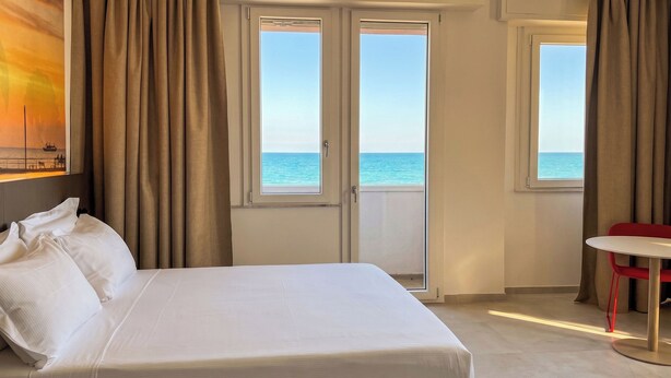 Habitación de hotel con cama, una mesa y vista al mar a través de grandes ventanales.