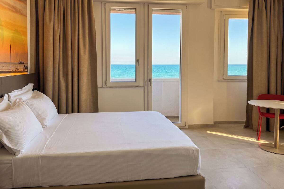 Une chambre d’hôtel avec un lit, une table et une vue sur la mer, grâce à de grandes fenêtres.