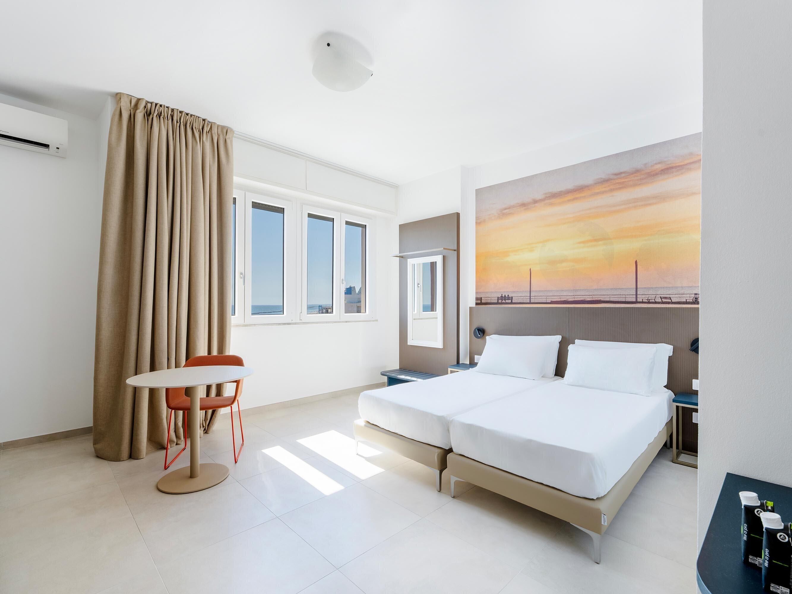 A hotel room with a bed, a table with a chair, a painting of a sunset, and a window with curtains.