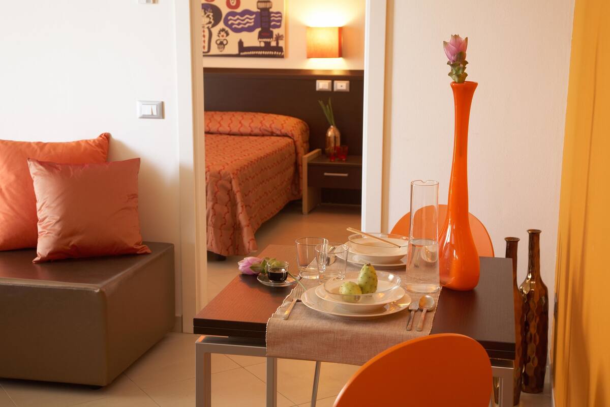 A hotel room with a dining area, a bed with orange bedding, a vase with a flower, and a table set for two.