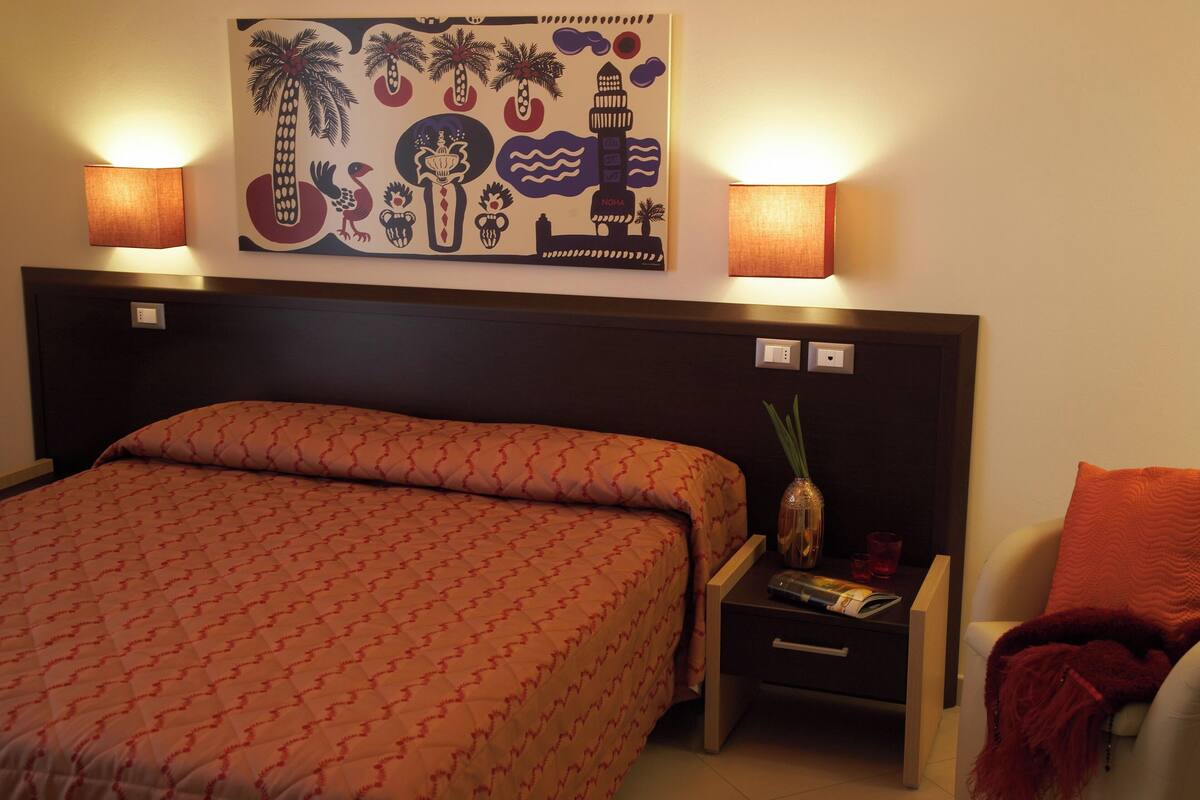 A hotel room with a bed, nightstands, a lamp, a wall art piece, and a chair.