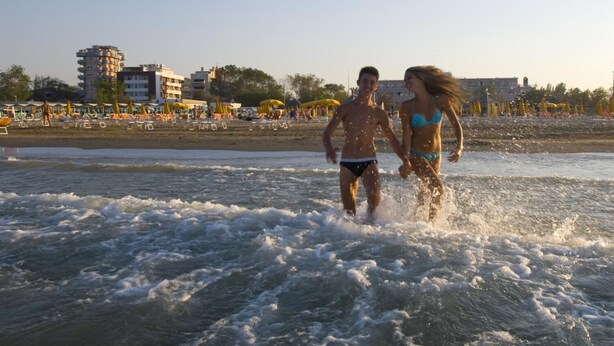 A beach with people running in the water, umbrellas, and buildings in the background. - Noha Suite Hotel (Riccione)
