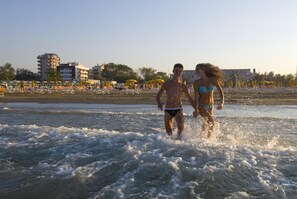 A beach with people running in the water, umbrellas, and buildings in the background. - Noha Suite Hotel (Riccione)