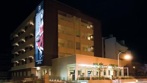 A modern hotel with multiple floors, a large billboard on the side, and a well-lit entrance area. - Noha Suite Hotel (Riccione)