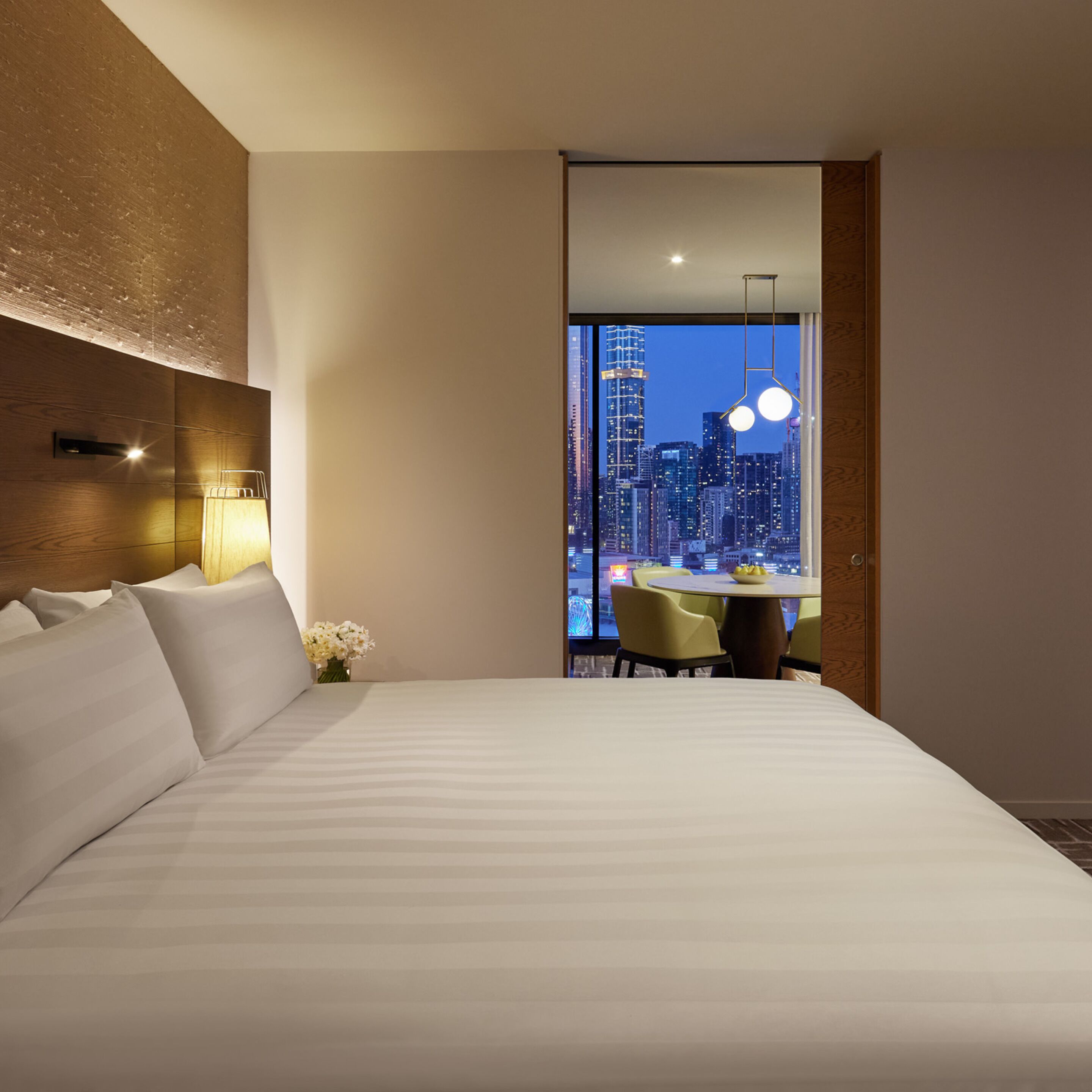 A modern hotel room with a large bed, a dining area with a table and chairs, and a cityscape view through a large window.