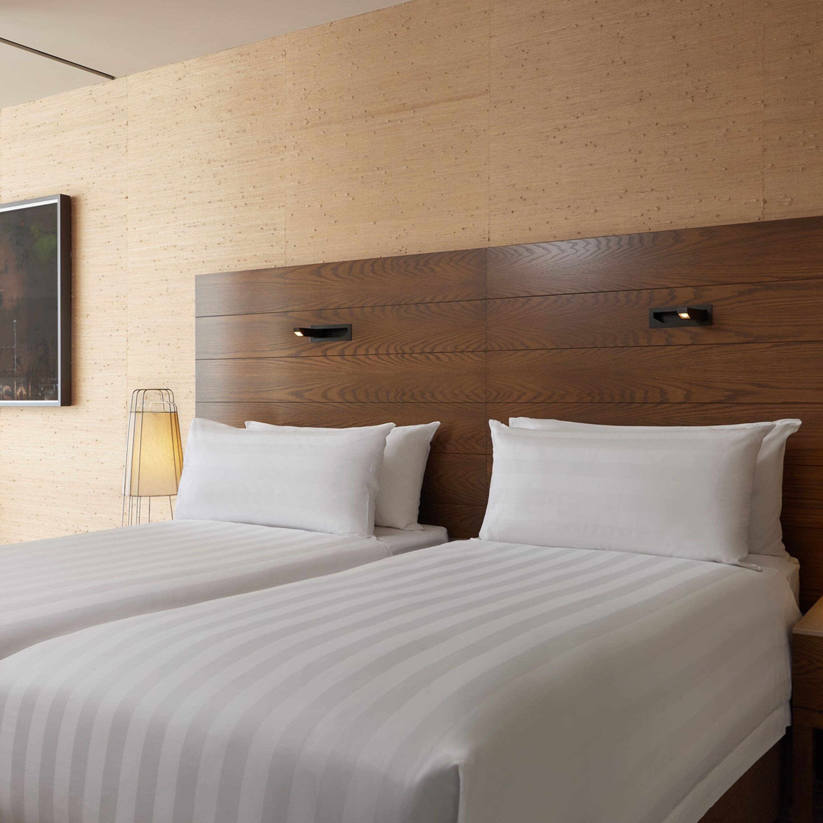 A hotel room with two beds, wooden headboards, a bedside lamp, and a framed picture on the wall.