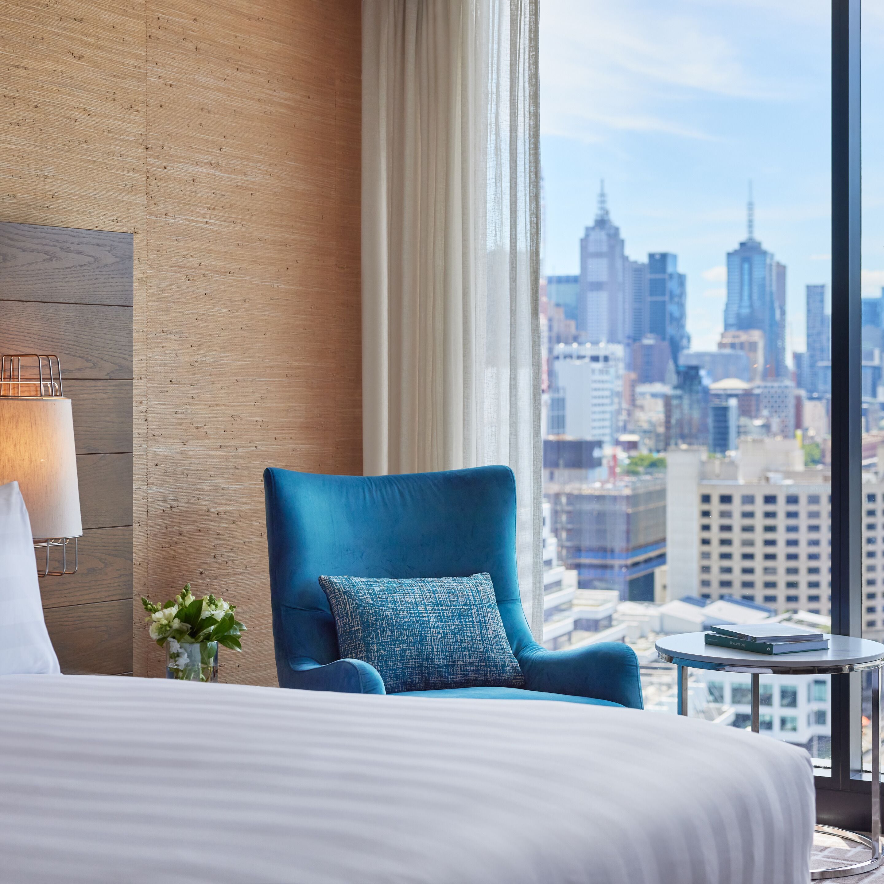 A hotel room with a large bed, a blue armchair, a small table, and a view of a city skyline.