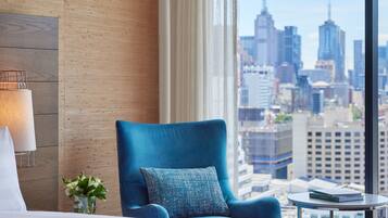 A hotel room with a large bed, a blue armchair, a small table, and a view of a city skyline.