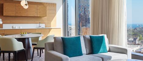 A modern living room with a grey sofa, teal cushions, a round coffee table, and a city view through large windows.
