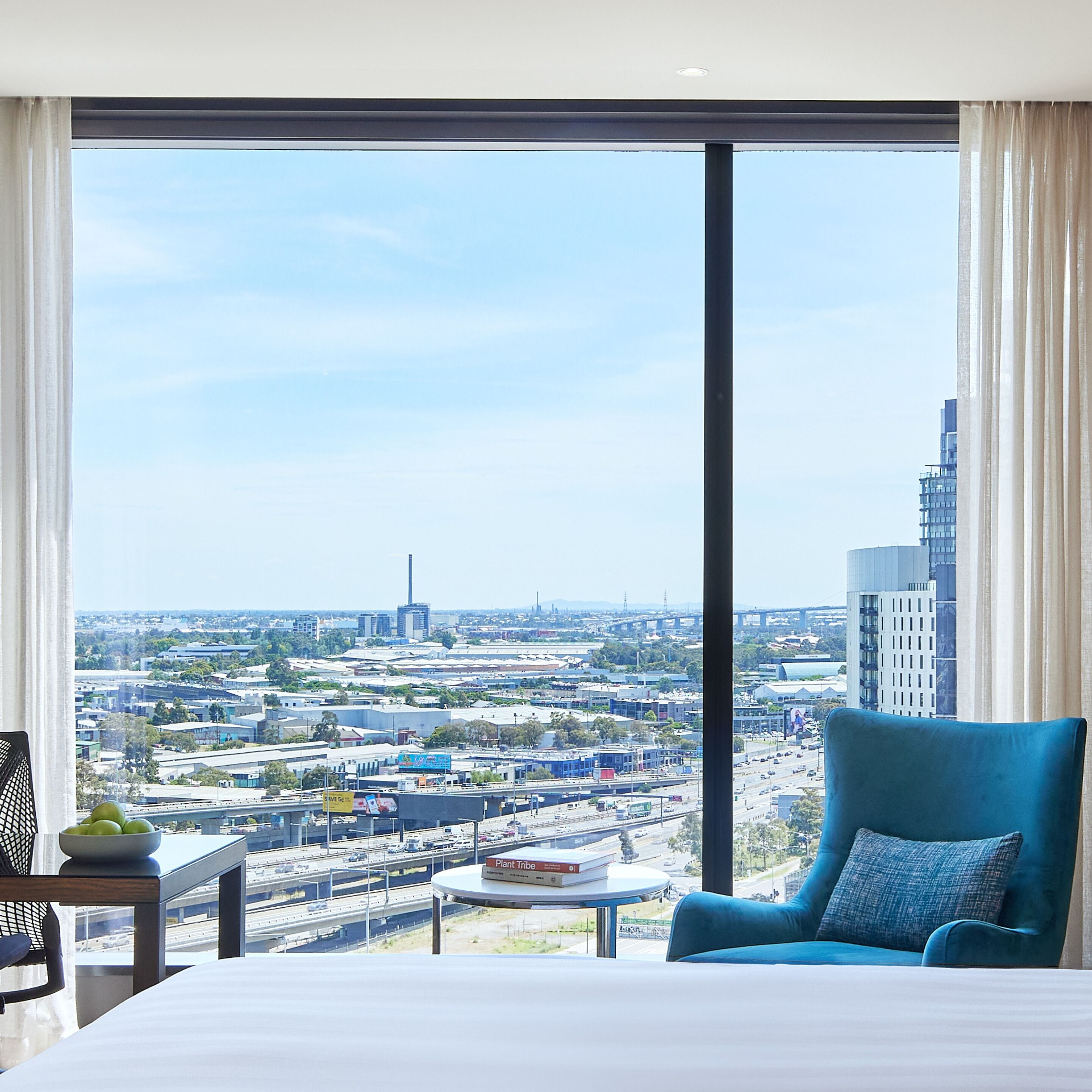 A hotel room with a large window offering a city view, a bed, a blue armchair, and a small table with a bowl of fruit and a book.