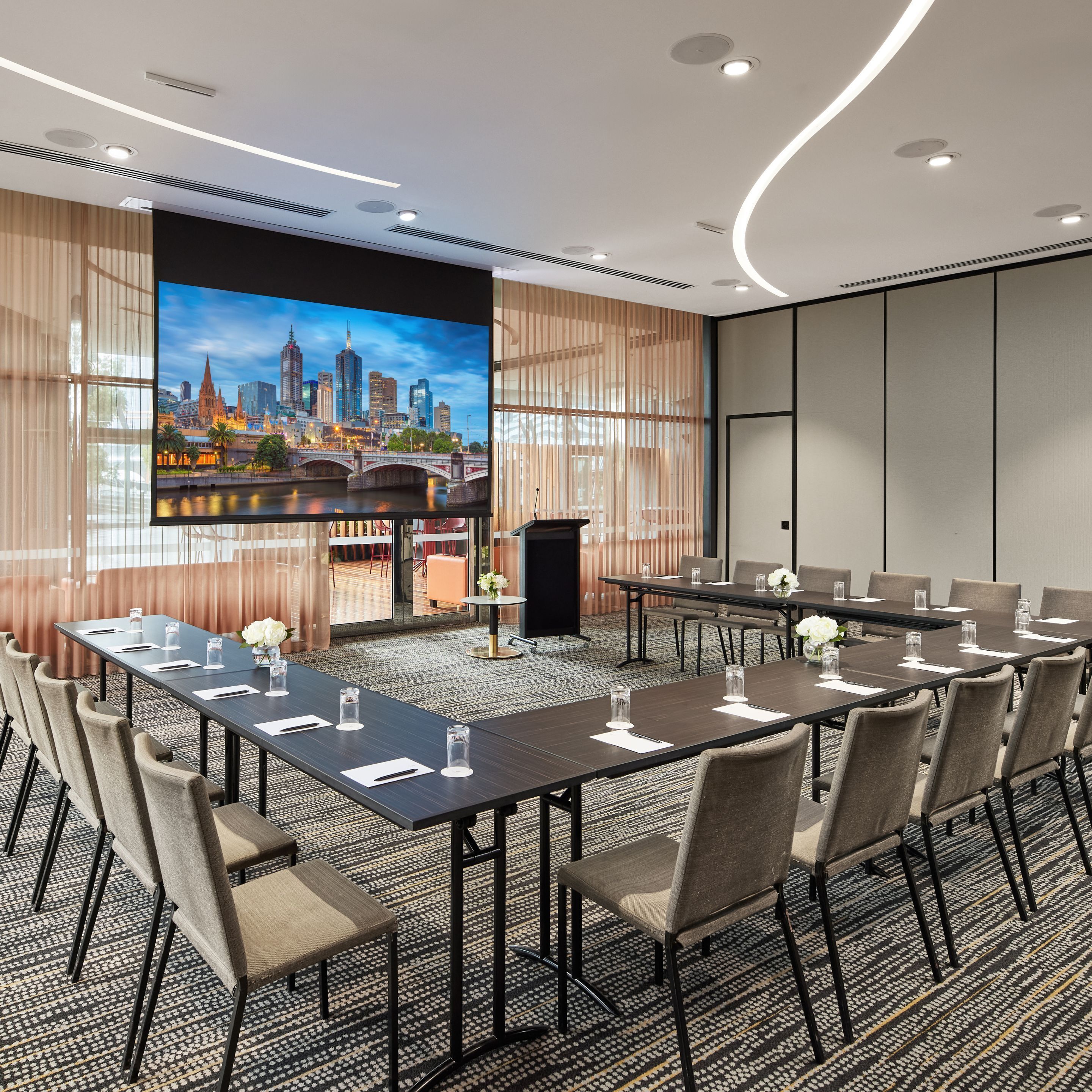 A conference room with a large screen displaying a cityscape, a long table with chairs, and a carpeted floor.
