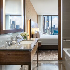 A modern hotel bathroom with a large mirror, dual sinks, a vanity, a bed with a city view, and a bathtub.