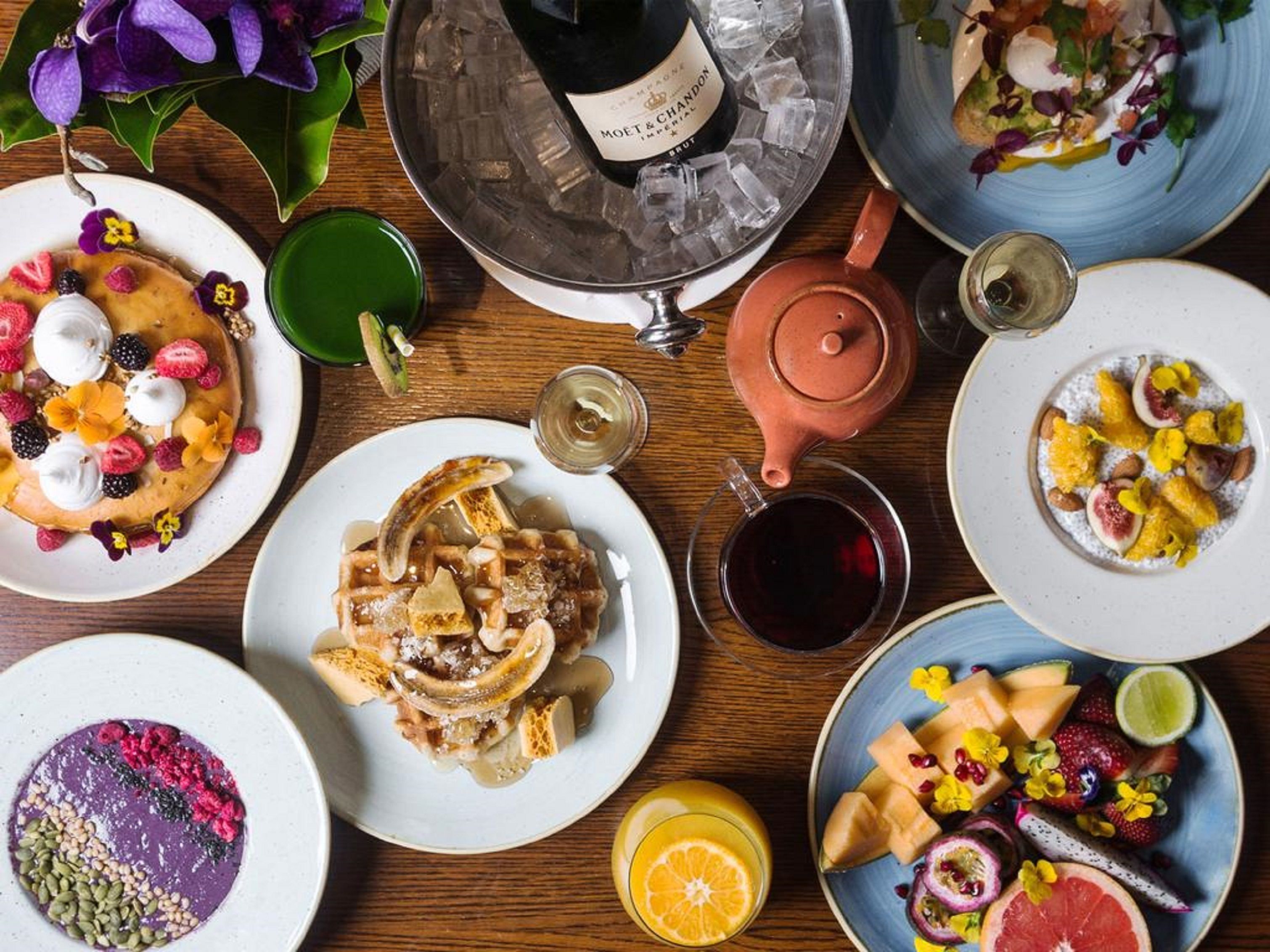 A variety of breakfast dishes, including pancakes, waffles, and fruit salads, served on a wooden table with a teapot and glasses of tea and juice.