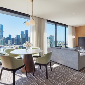 A modern hotel room with a city view, a round dining table, green chairs, a sofa, and a flat-screen TV.