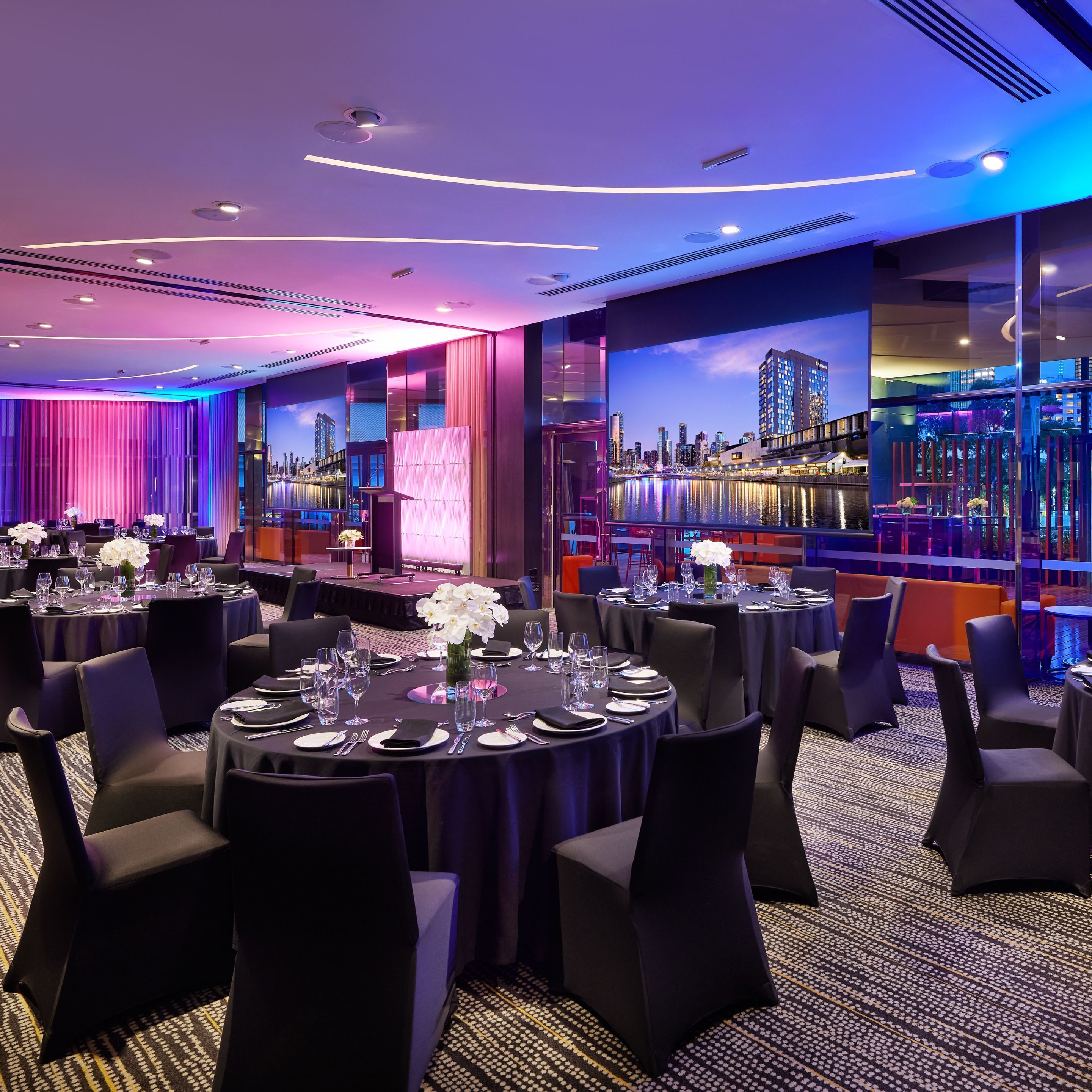 A banquet hall with round tables set for dining, featuring a cityscape view through large windows.