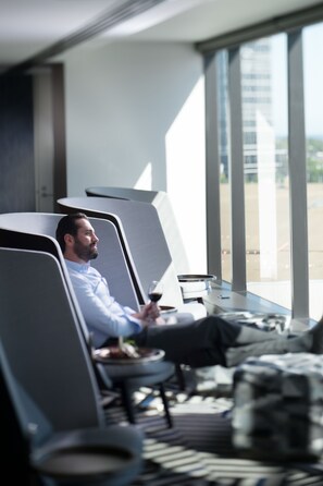 A man reclining in a modern lounge chair with a drink, large windows, and a view of a building.