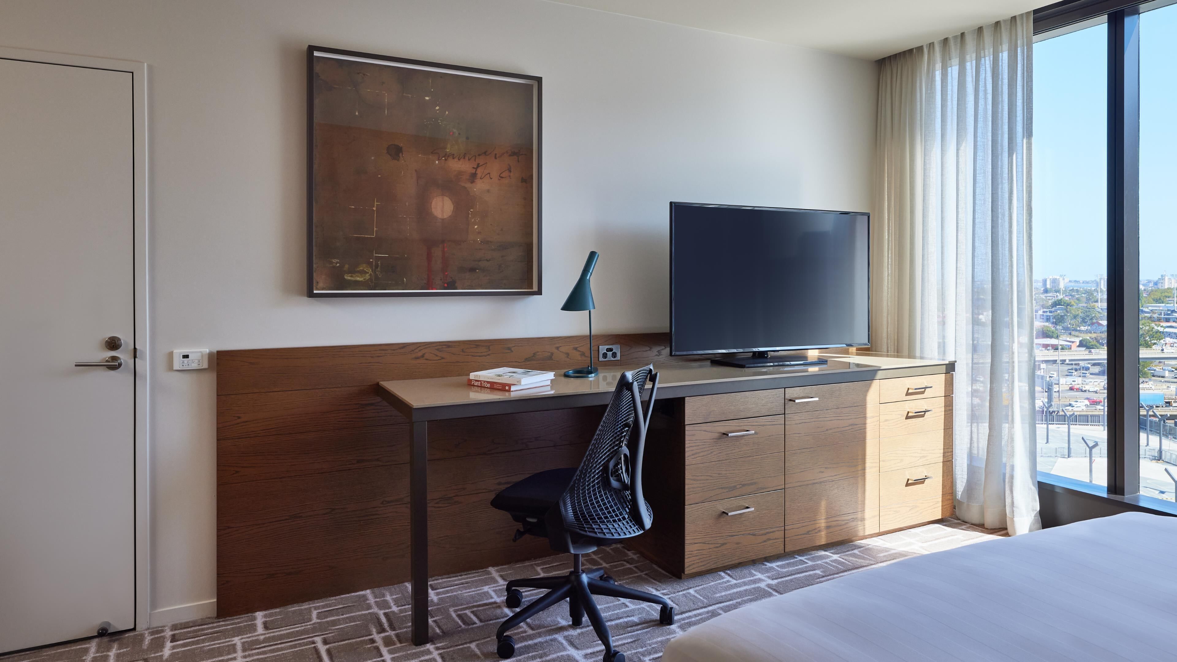 A hotel room with a bed, a desk with a TV, a chair, a painting, and a view of the city.