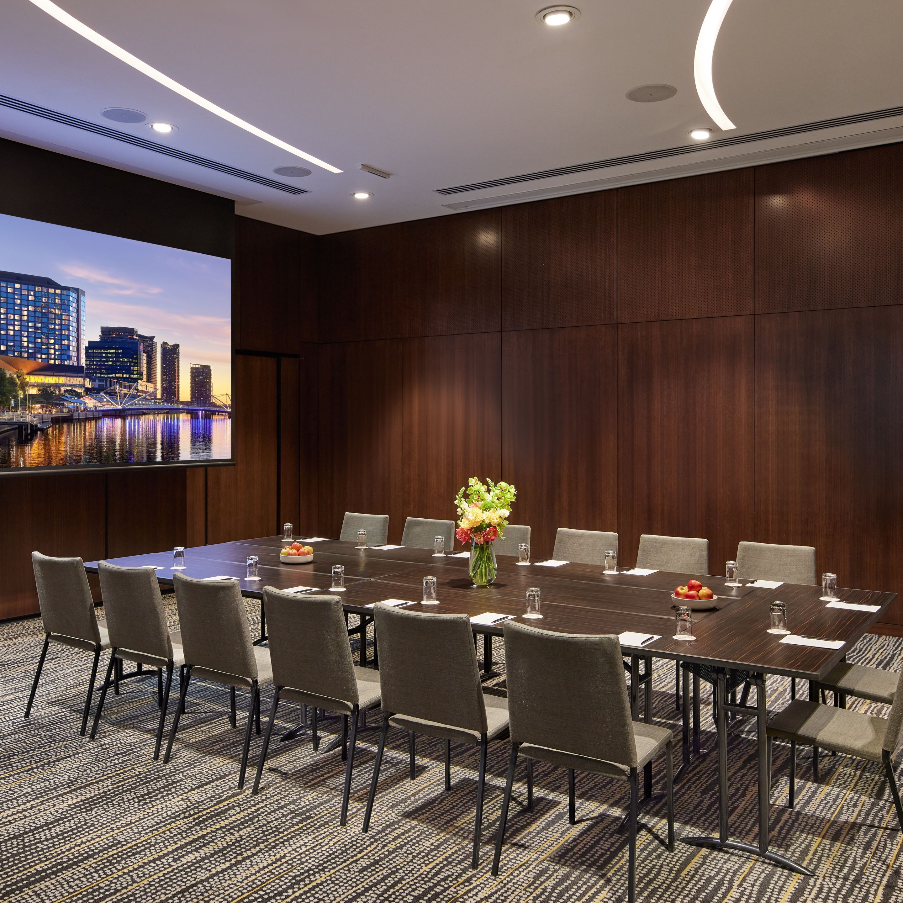 Un salón de conferencias con una gran mesa, sillas y una pantalla de proyector que muestra un paisaje urbano.