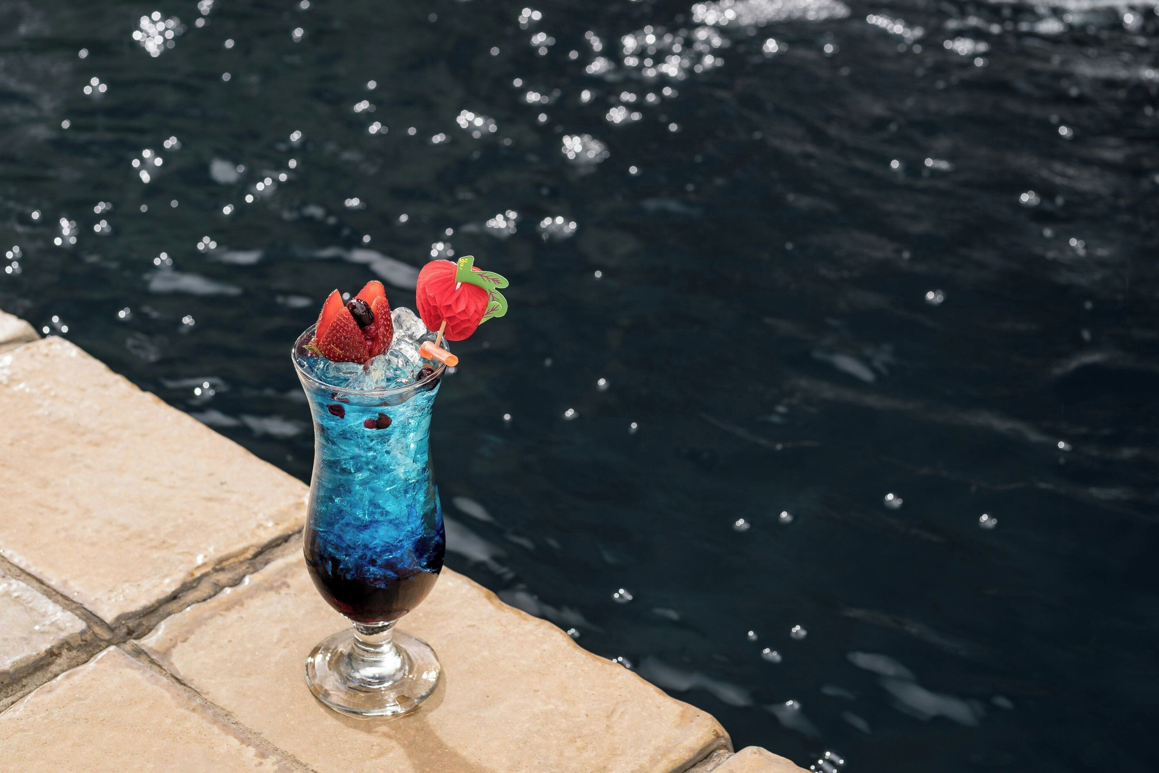 A glass of blue cocktail with strawberries on a stone ledge by water.