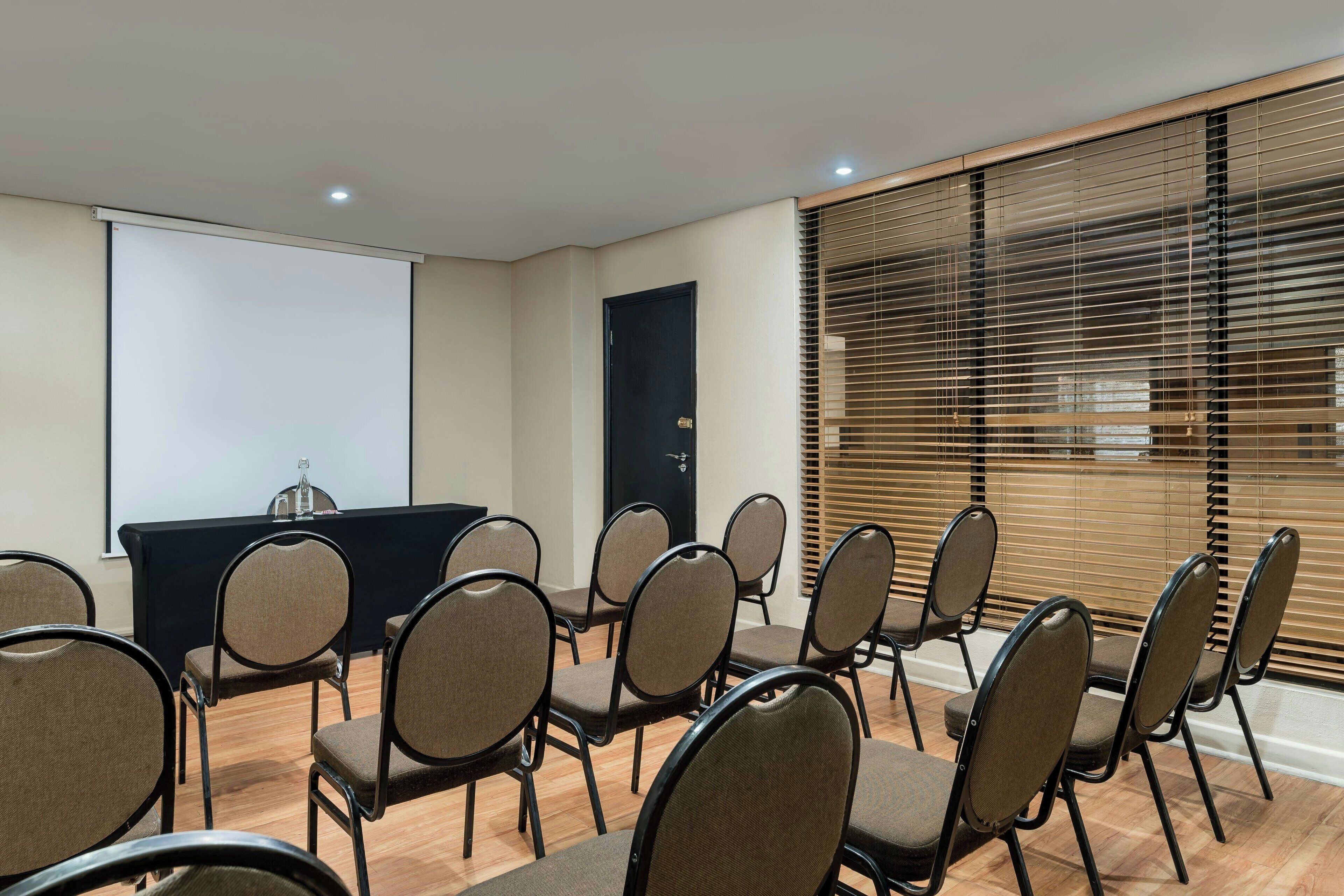 A conference room with a projector screen, a black table with a water bottle, and rows of chairs.