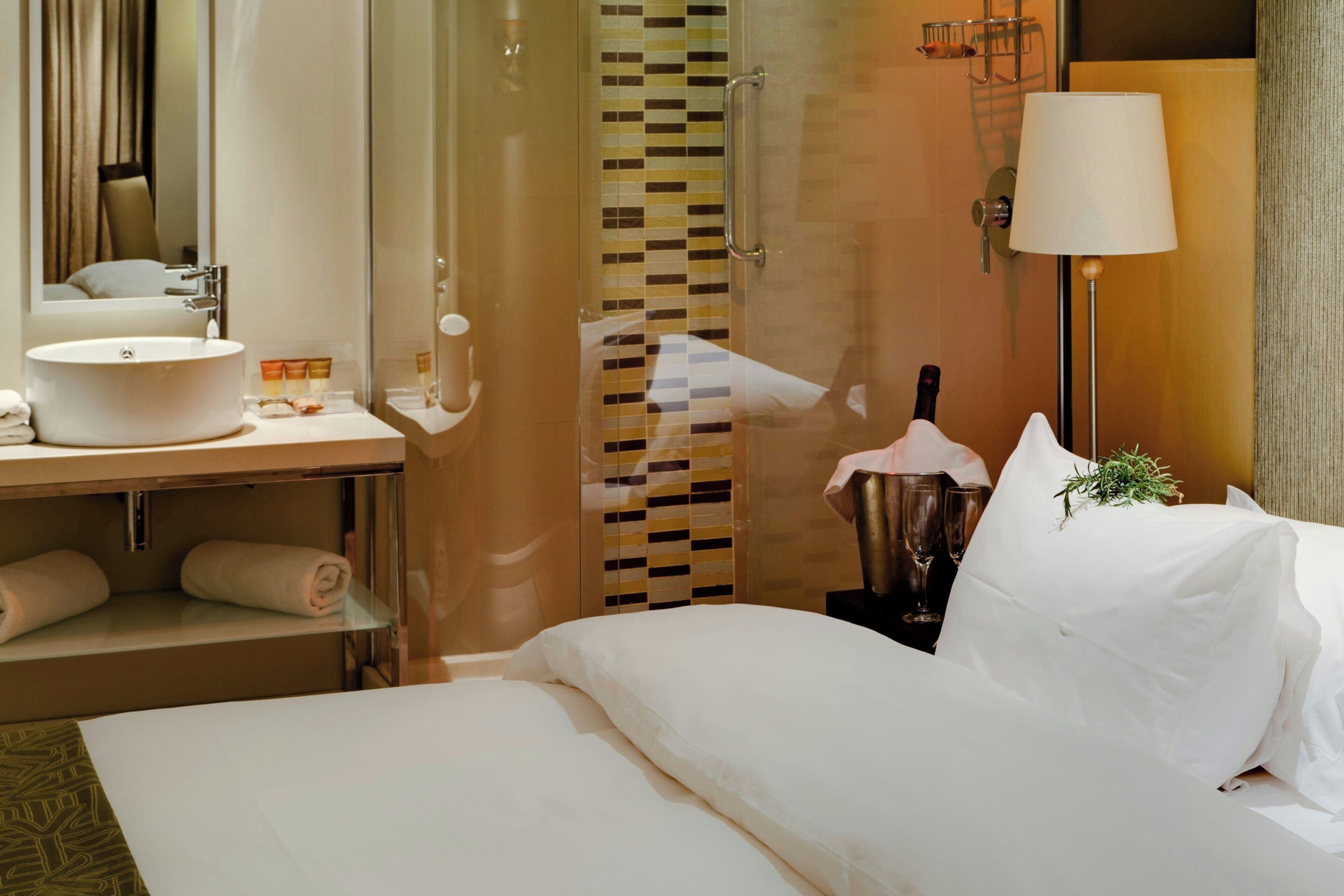 Une chambre d’hôtel avec un grand lit, une table de chevet avec une lampe, une salle de bain avec un lavabo et un miroir, et un espace douche.