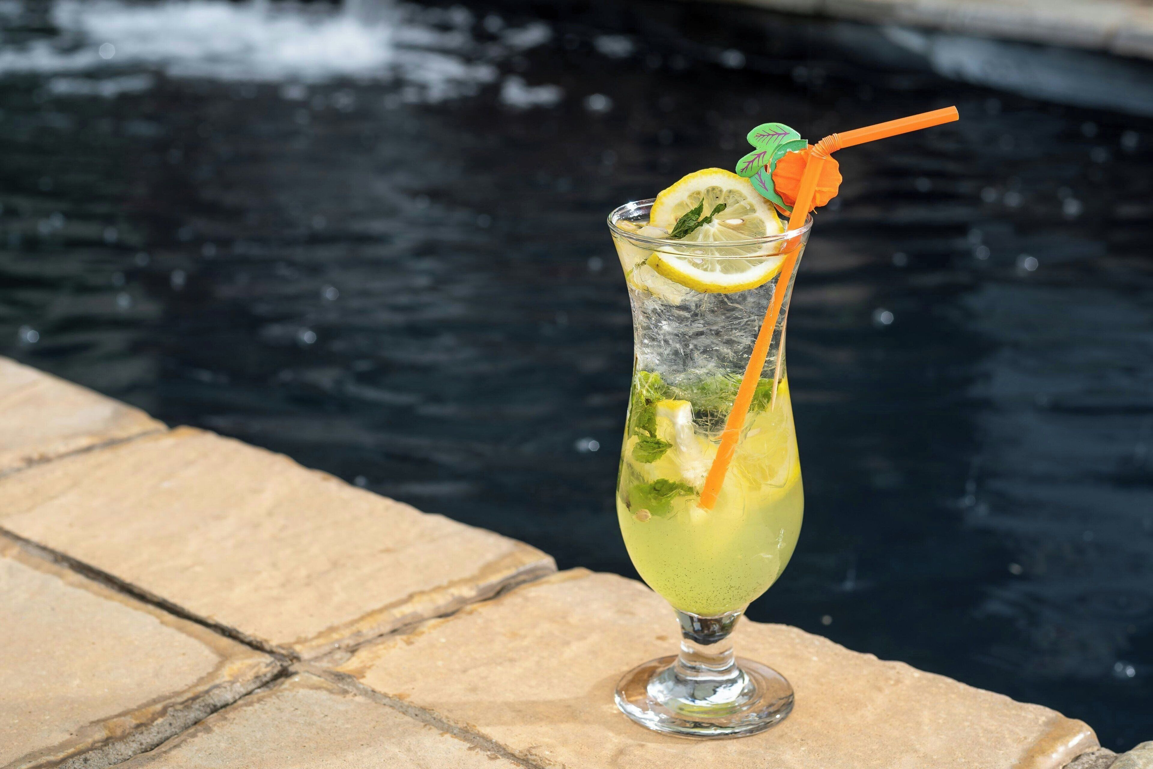 A cocktail with a lemon slice and mint garnish, placed on a stone ledge by a body of water.
