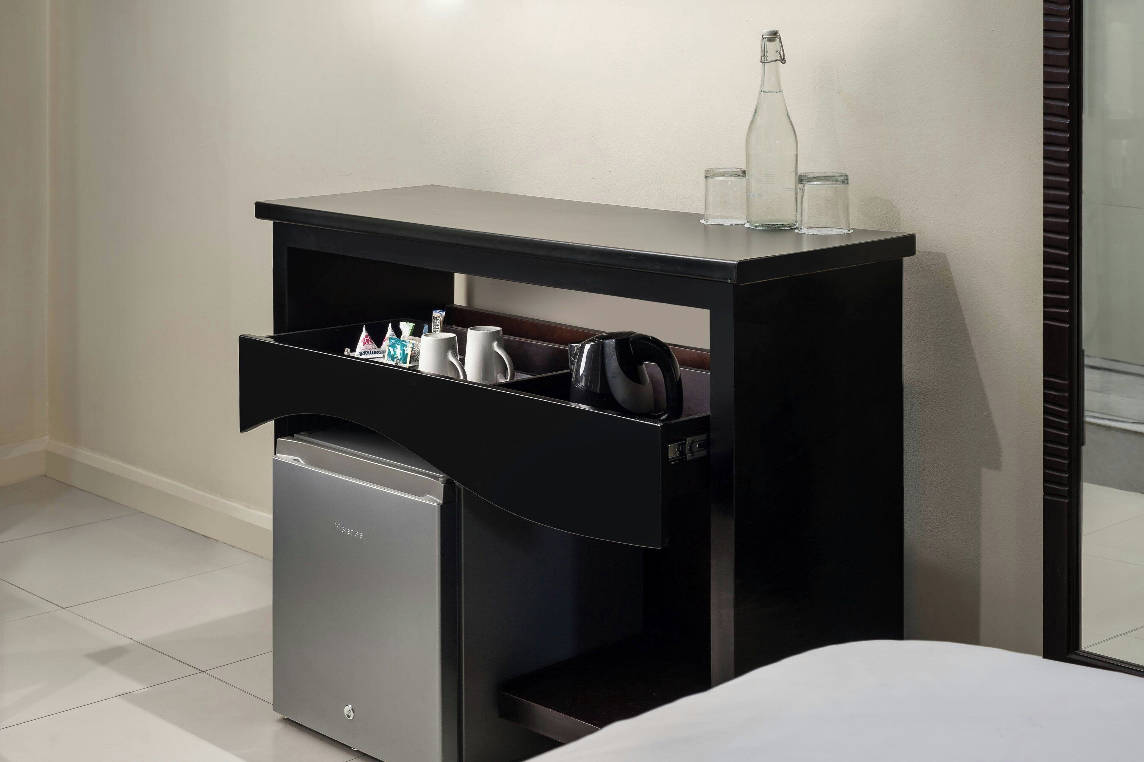 A compact hotel room with a small desk featuring a drawer, a compact refrigerator, and a glass bottle on the desk.
