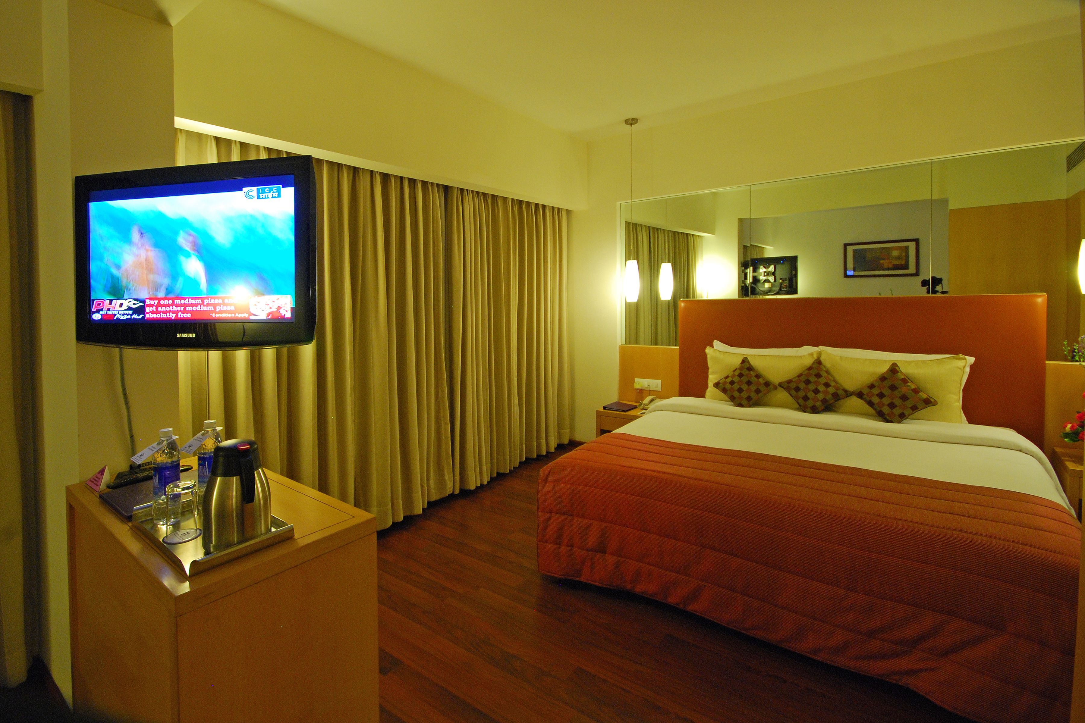 A hotel room with a bed, a TV on a stand, a desk with a laptop, and a mirror.