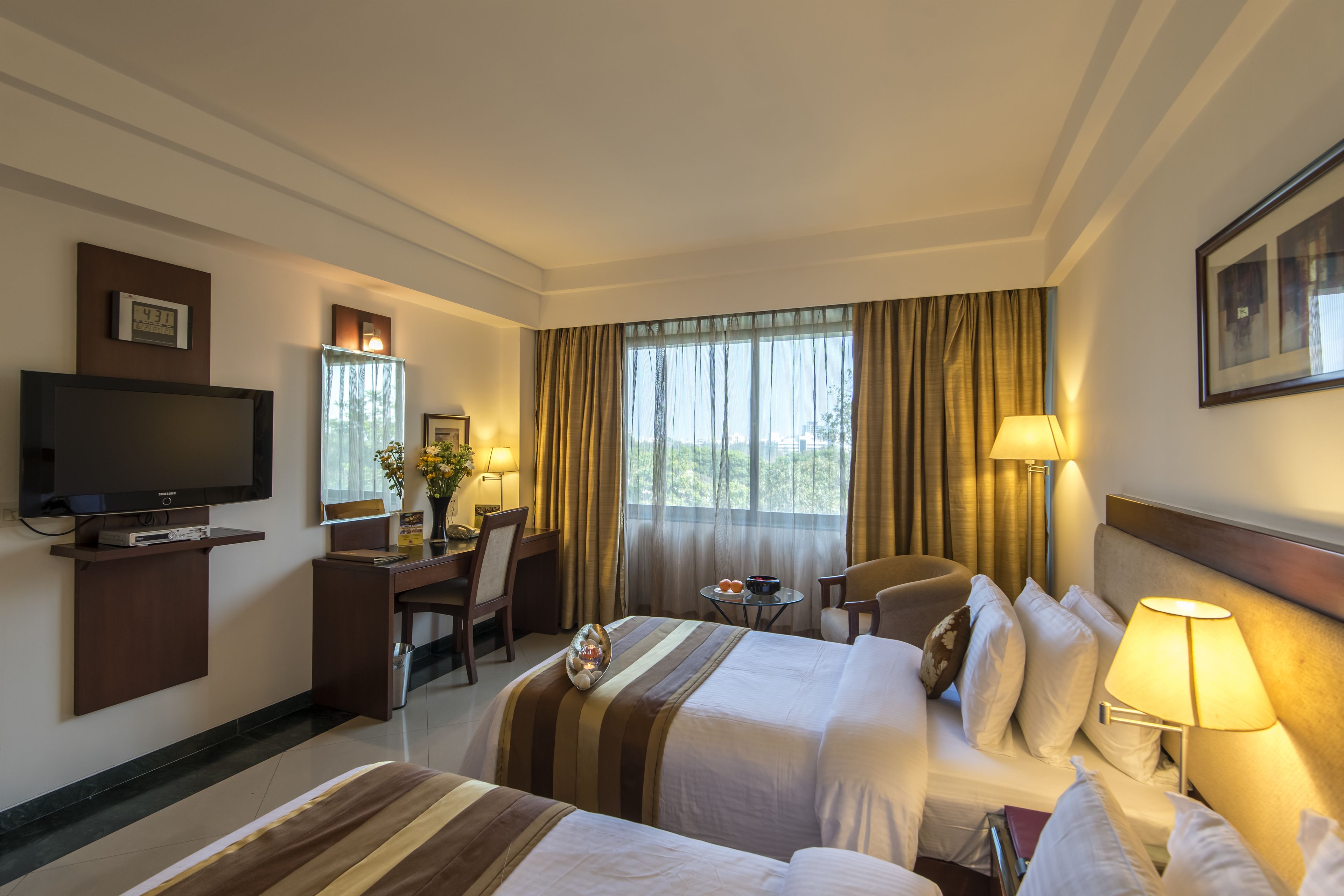 A hotel room with two beds, a desk with a television, a chair, a small table with a lamp, and a window with curtains.