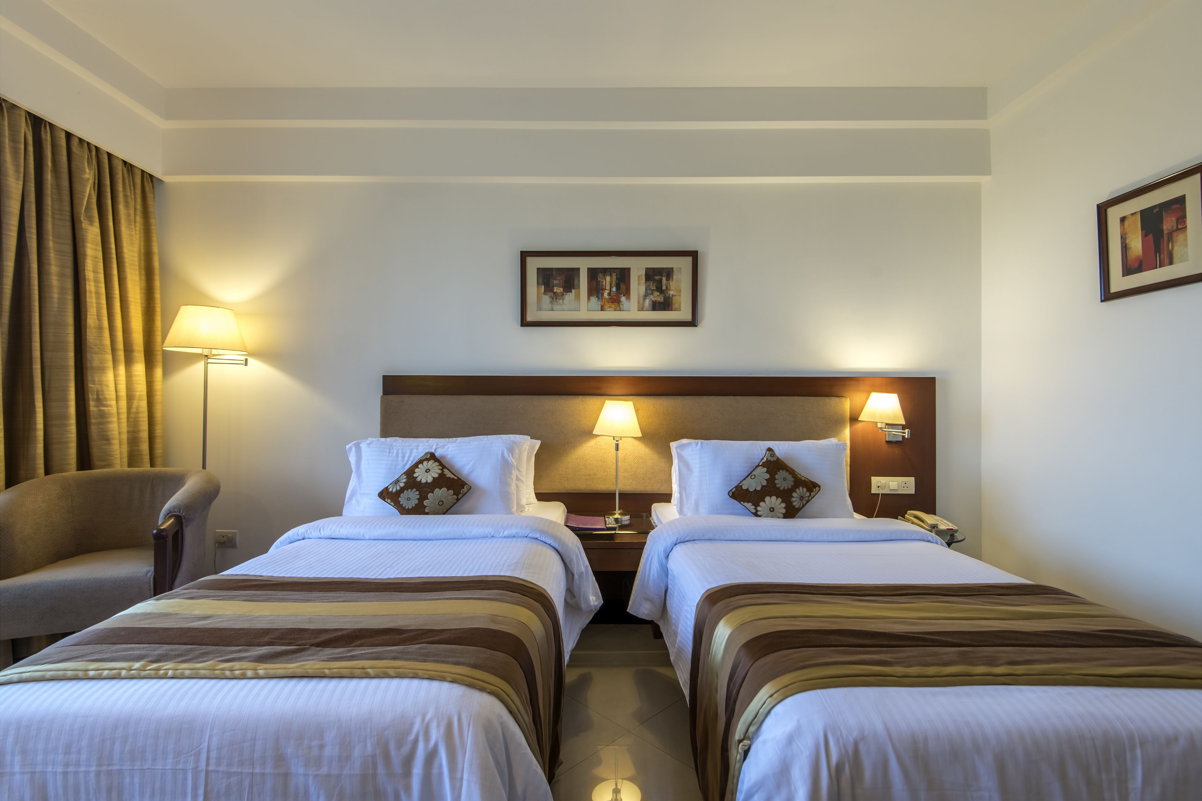 A hotel room with two beds, a chair, a nightstand with a lamp, and a framed picture on the wall.