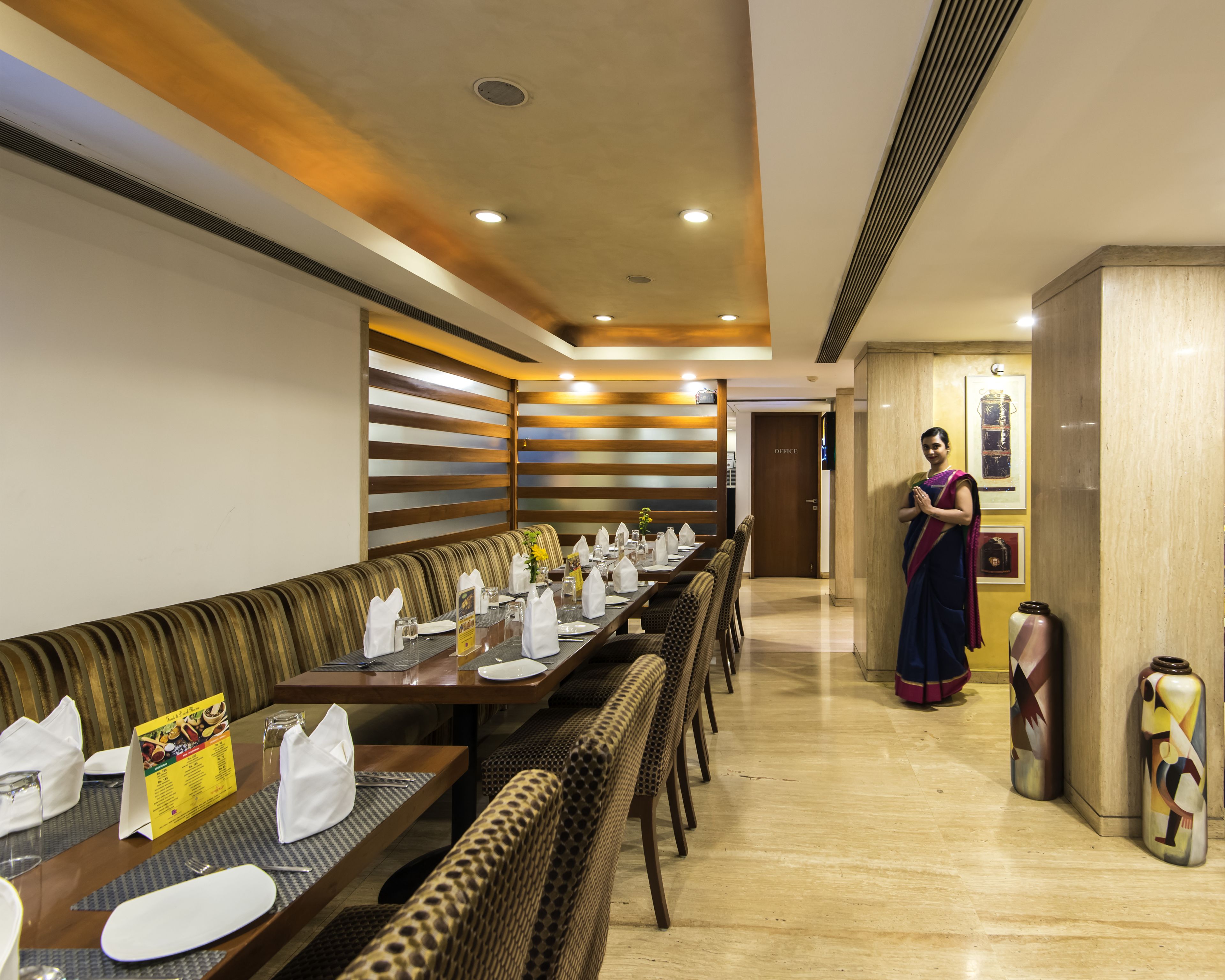 Ein Restaurant mit einem langen Esstisch, der für eine Mahlzeit gedeckt ist. Im Hintergrund steht eine Frau an der Wand und eine Tür mit der Aufschrift 'OFFICE'.