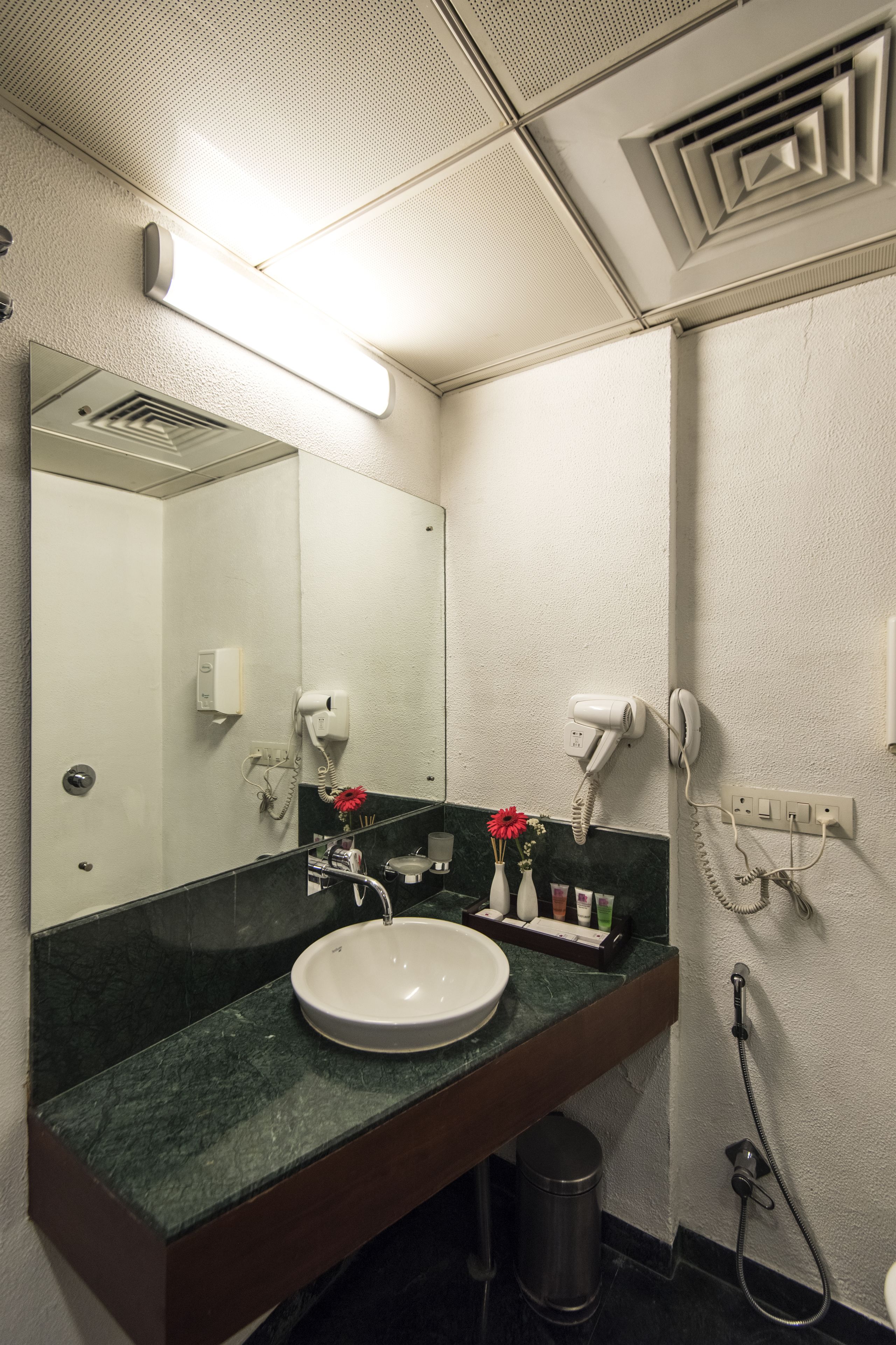 A bathroom with a white sink, dark countertop, and a mirror. There are hair dryers and a phone on the wall.