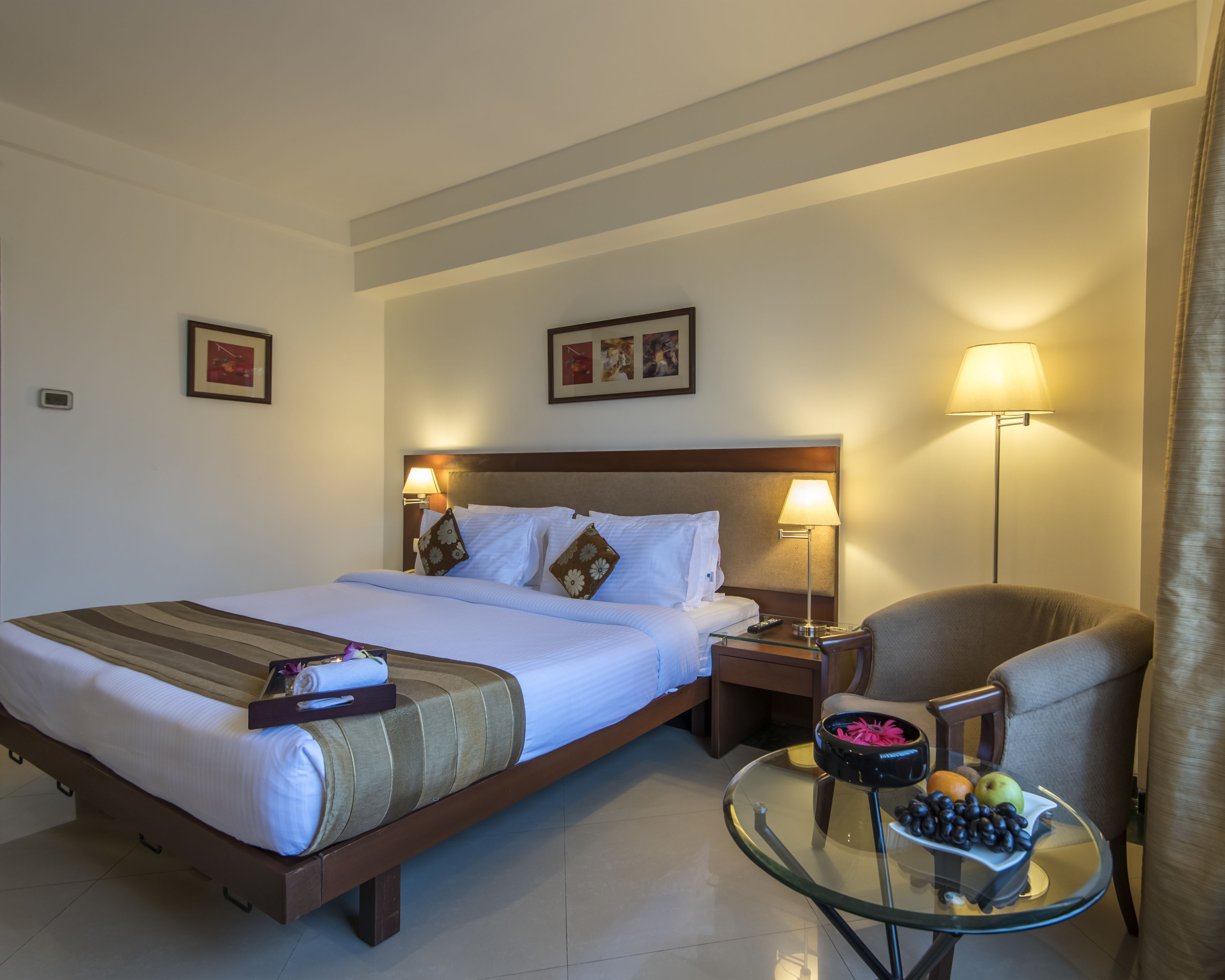 Una habitación de hotel con una cama grande, dos lámparas de noche, una mesita de vidrio con frutas y una silla.