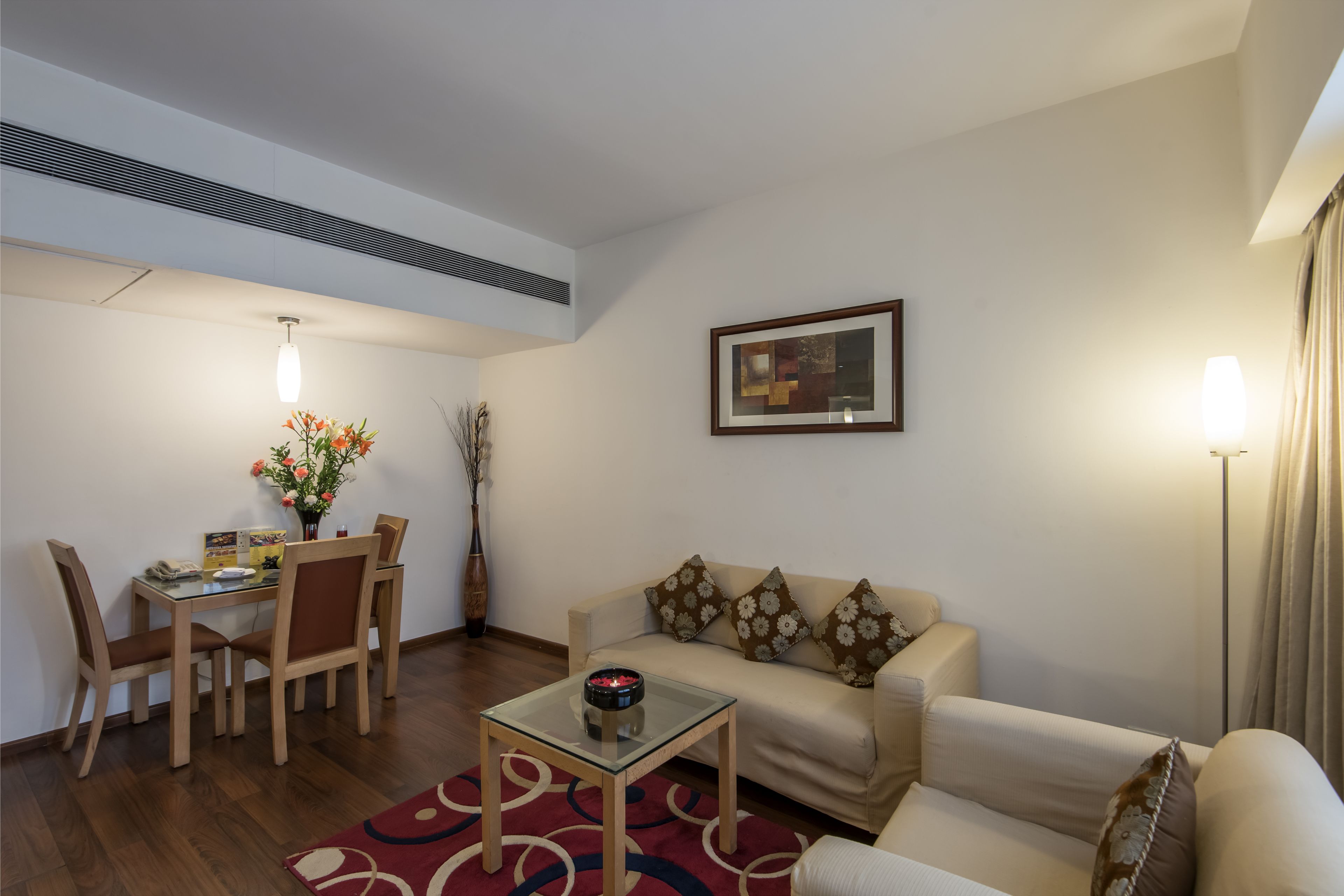 A living room with a sofa, a glass coffee table, a dining table with chairs, and a vase with flowers.