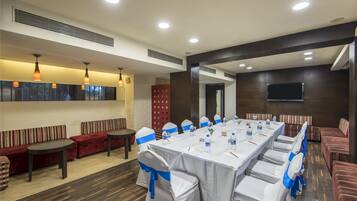 Un salón de conferencias con una larga mesa, sillas blancas adornadas con cintas azules y un televisor instalado en la pared.
