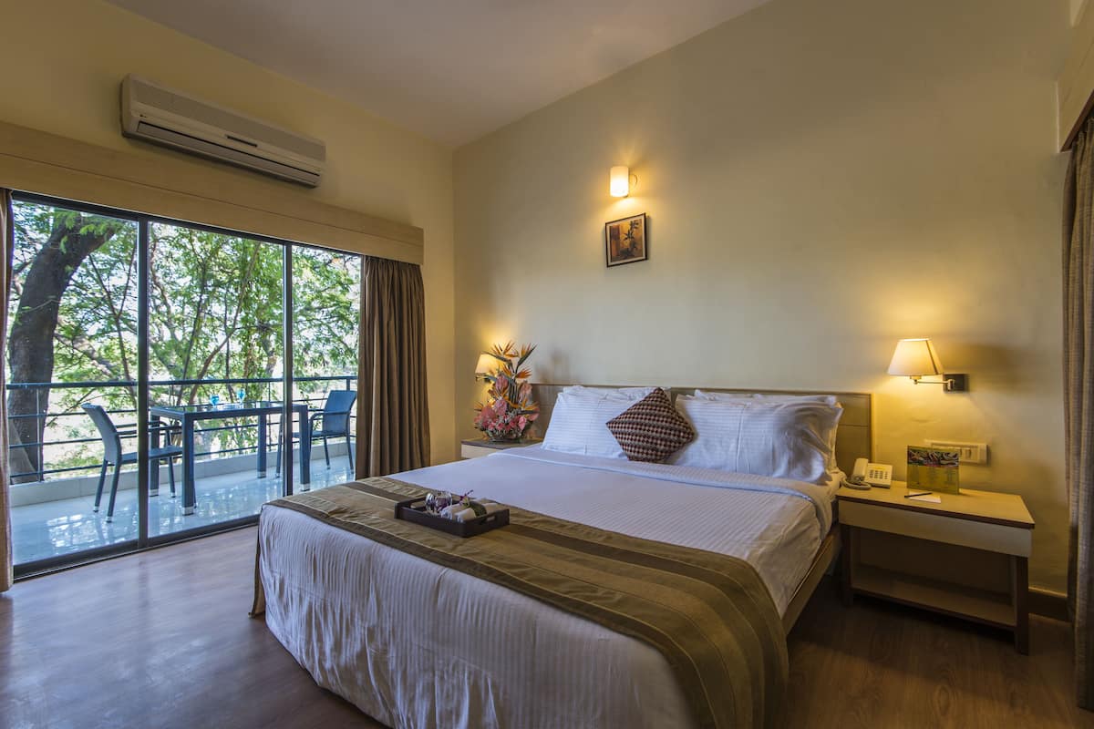 A hotel room with a large bed, a bedside table with a lamp, a nightstand with a phone, a window with a view of trees, and a balcony with chairs.