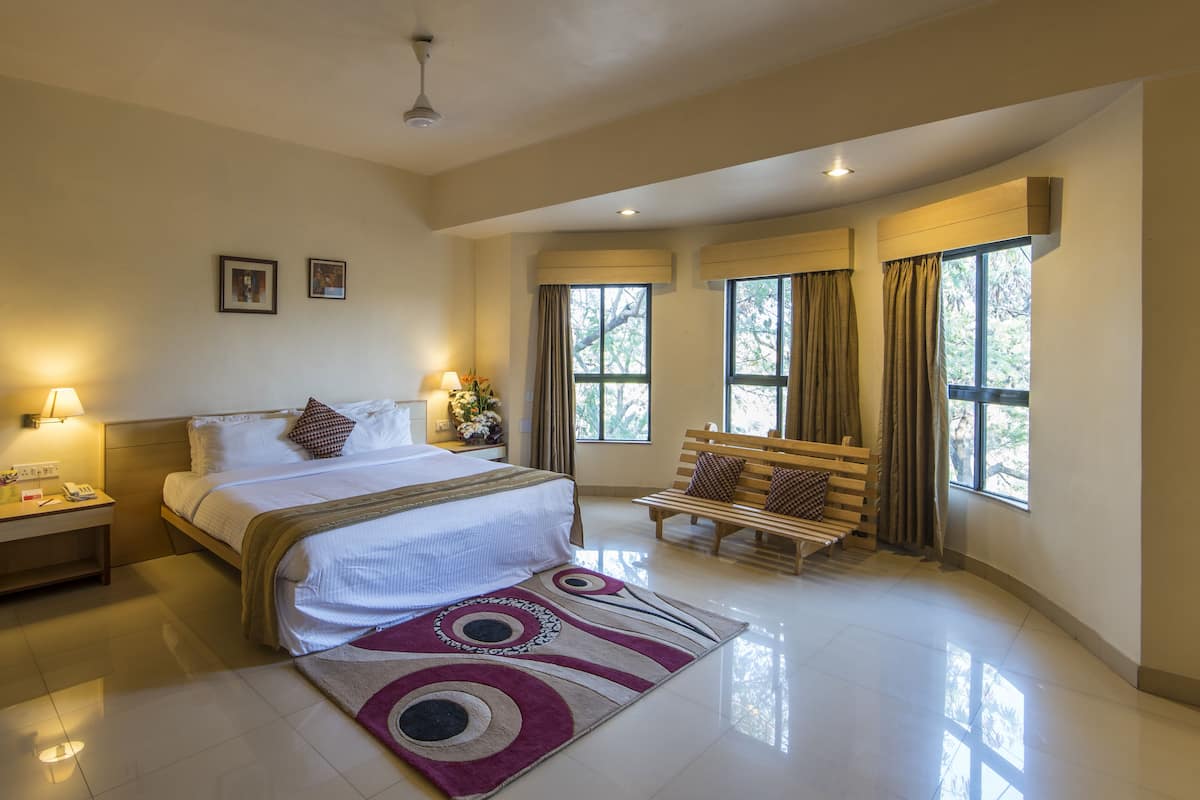 A spacious bedroom with a large bed, bedside lamps, a bench, and a view of trees through the windows.