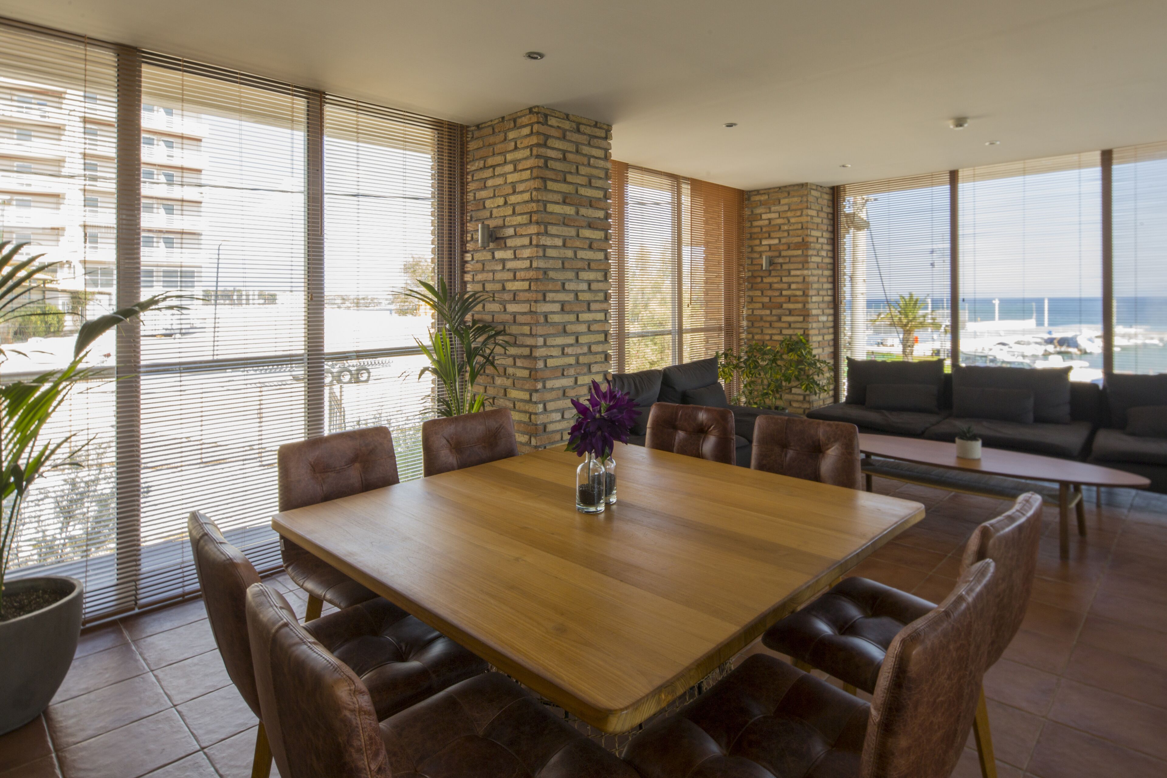 A spacious dining area with a wooden table and leather chairs, large windows with blinds, and a view of the sea.