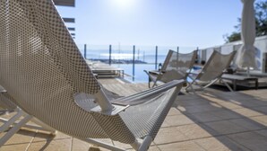 Uma área junto à piscina com espreguiçadeiras e guarda-sóis, céu claro e vista para o mar.