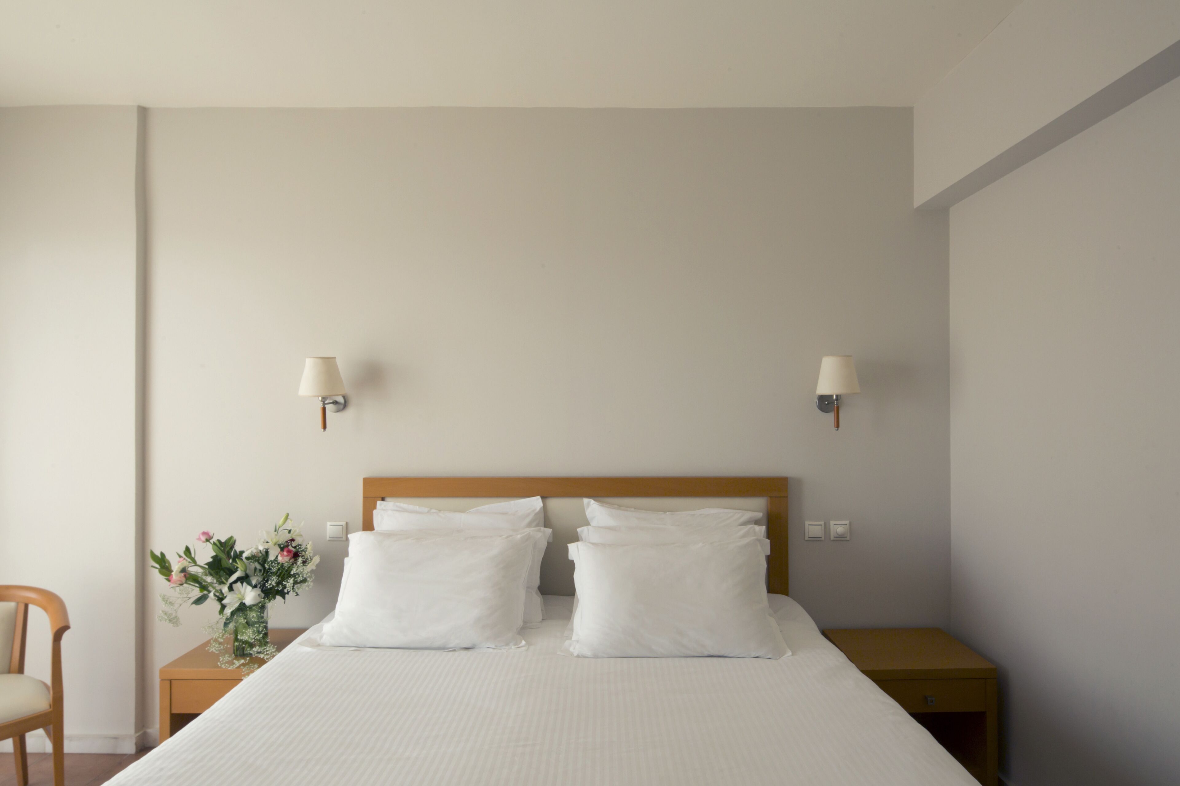 Una cama bien hecha con sábanas y almohadas blancas, una mesita de noche de madera con un florero y dos lámparas fijadas a la pared.