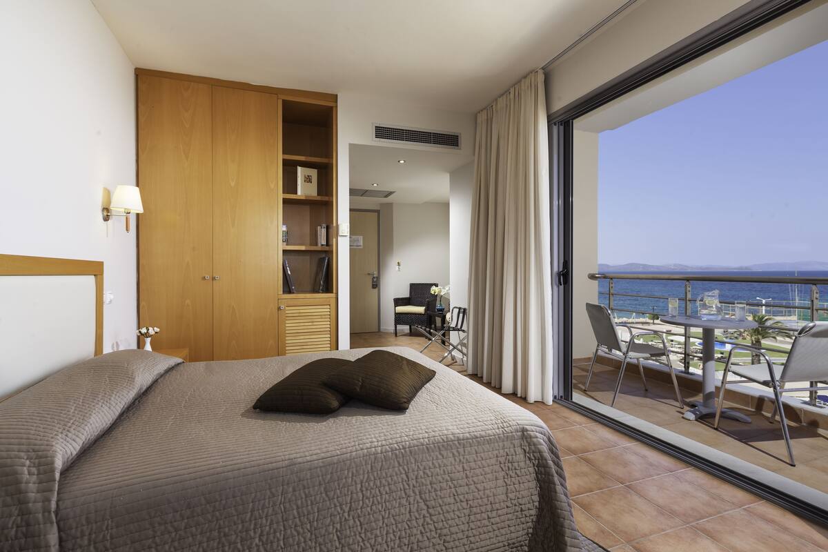 A hotel room with a bed, a wardrobe, a desk with a chair, and a balcony with a view of the sea.