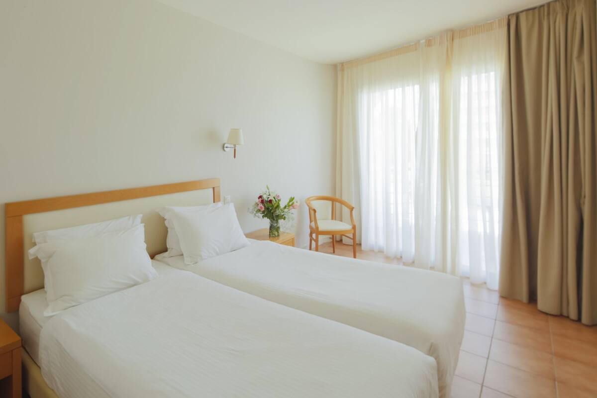A hotel room with two beds, a nightstand, a chair, a lamp, and a window with curtains.