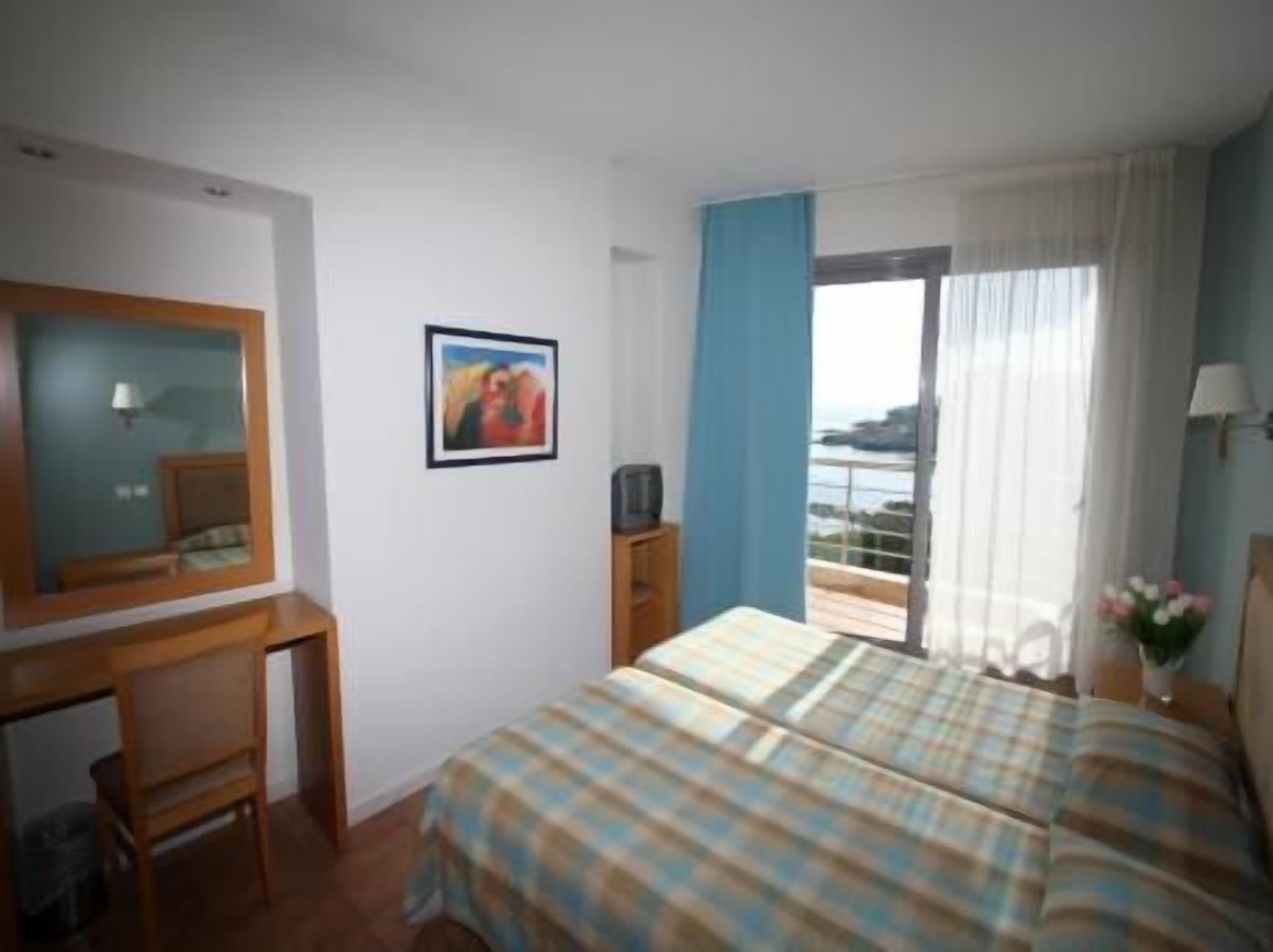 Habitación de hotel con cama, escritorio, silla, televisor, un cuadro en la pared y balcón con vistas.