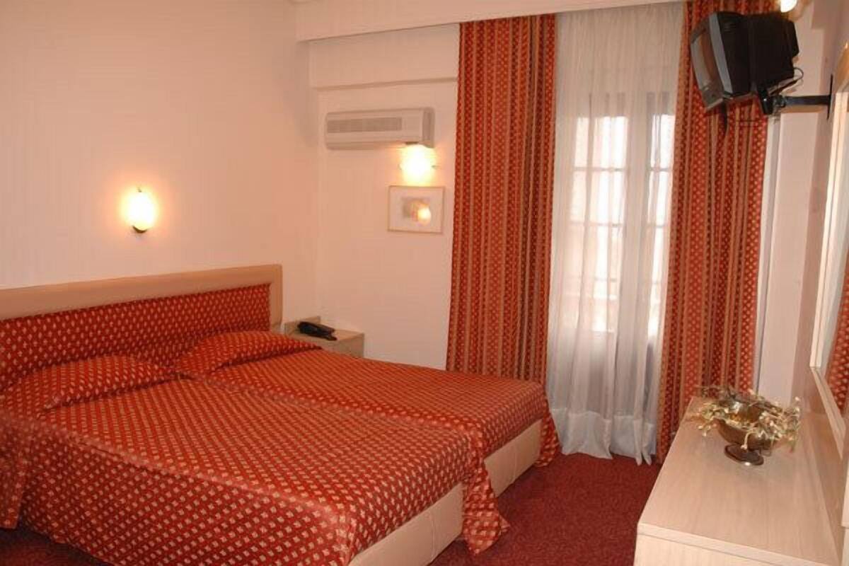 Habitación de hotel con cama doble, mesita de noche con lámpara, televisión montada en la pared y ventana con cortinas.
