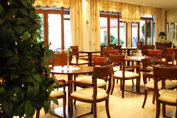 A hotel dining area with multiple wooden tables and chairs, large windows with curtains, and a potted plant in the foreground.