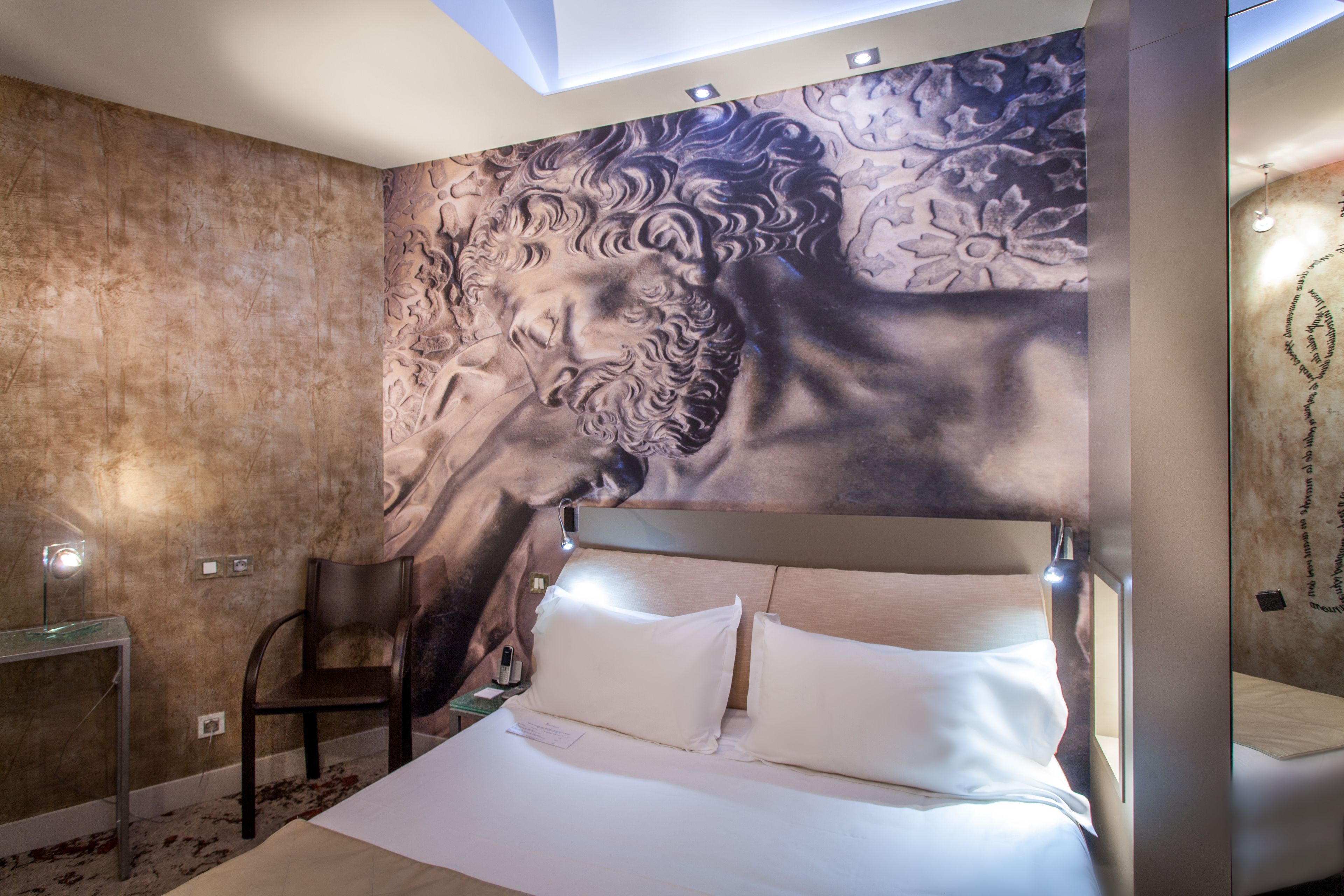 Quarto de hotel moderno com um grande mural, uma cama com roupa de cama branca, uma cabeceira de madeira e uma pequena mesa com uma cadeira.