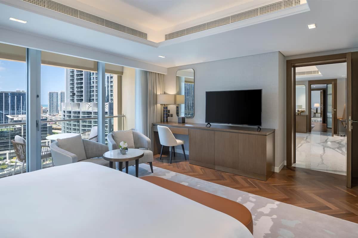 A modern hotel room with a large bed, a TV, a desk, and a balcony with a city view.