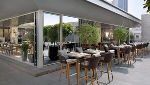 An outdoor dining area with tables and chairs, large potted plants, and a glass wall.